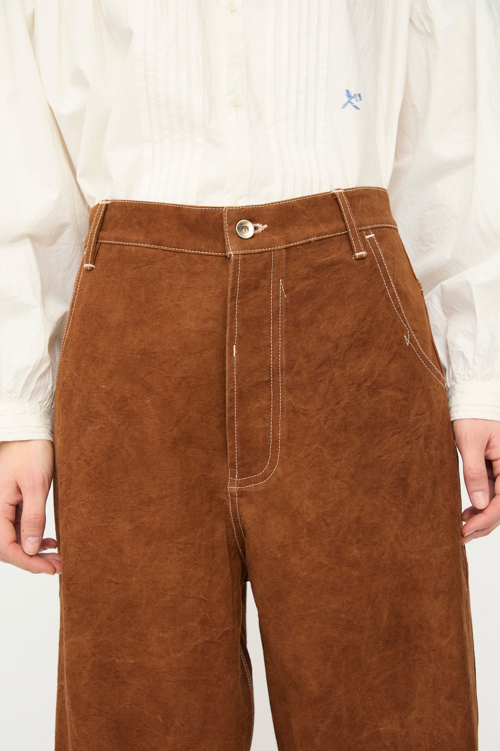 A person wearing Story mfg.'s Canvas Home Pant in Brown Wonky-Wear, paired with a white button-up shirt with pintuck detailing.