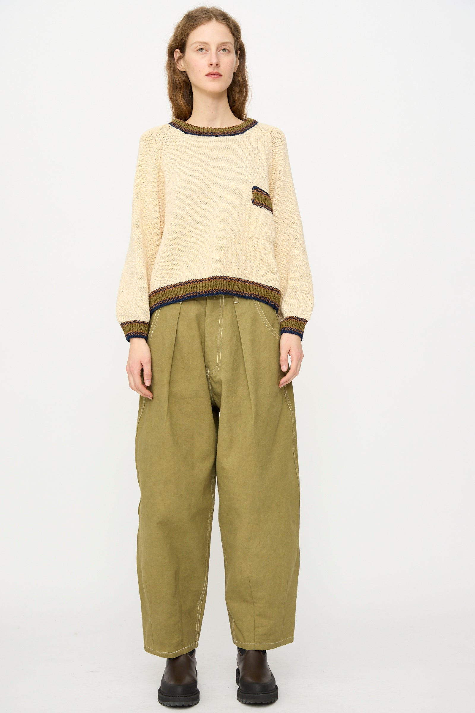 A person stands against a plain background wearing a beige sweater, Story mfg.'s Canvas Lush Pant in Olive Wonky-Wear, and black boots.