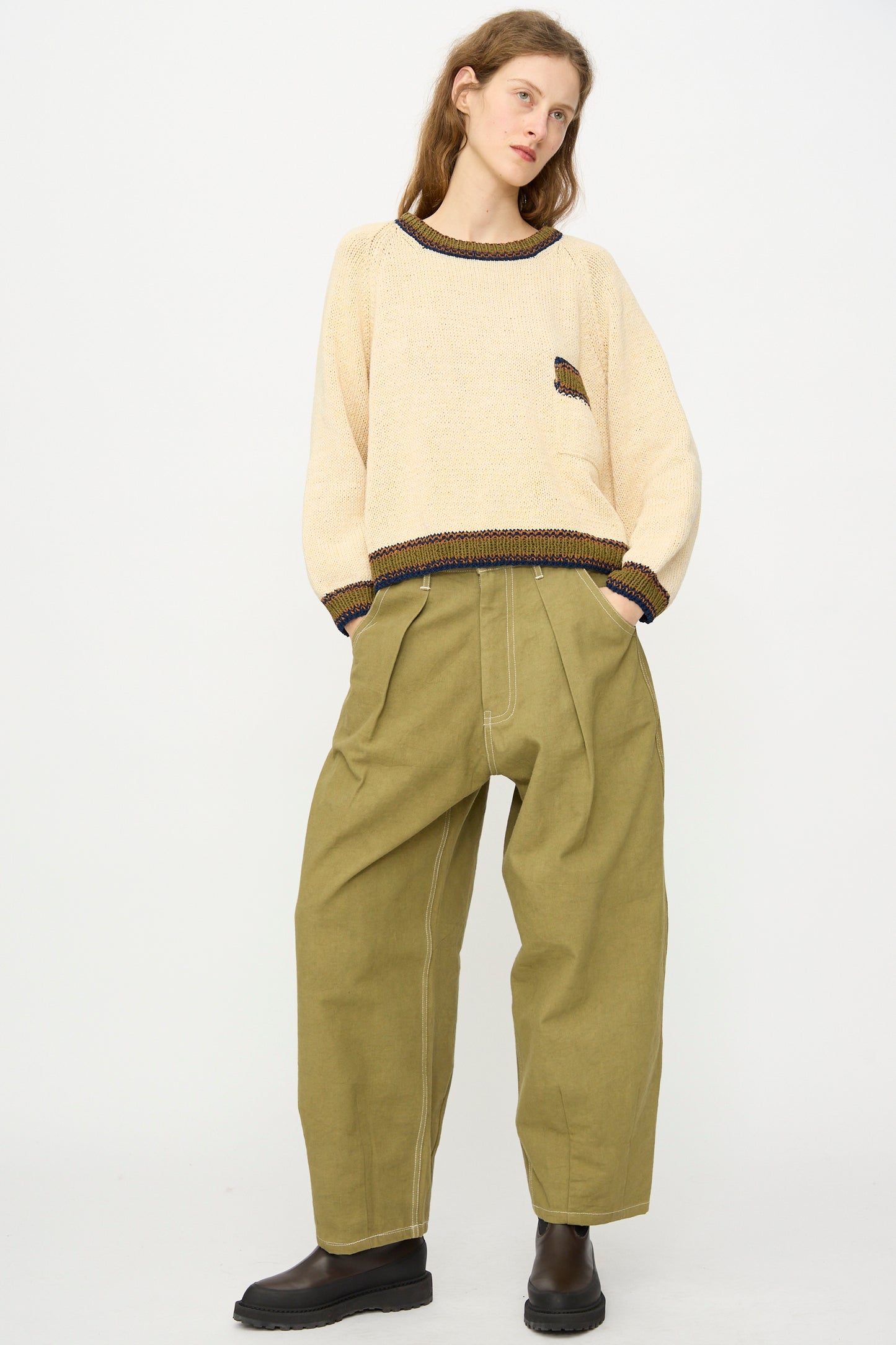 A person stands against a plain background wearing a beige sweater, Story mfg.'s Canvas Lush Pant in Olive Wonky-Wear with visible seams, and black shoes.