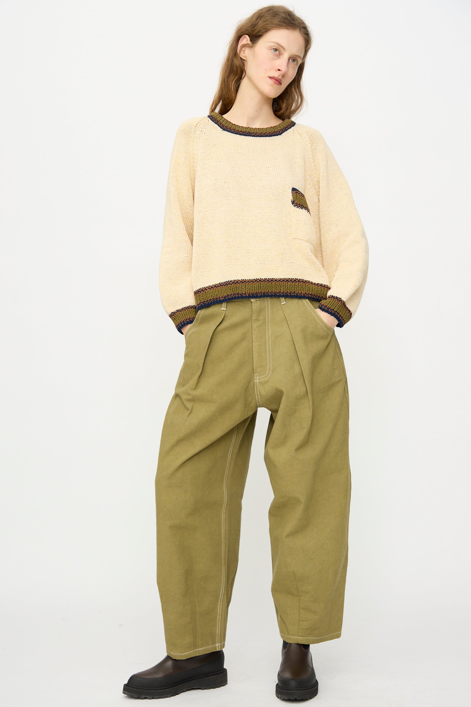 A person stands against a plain background wearing a beige sweater, Story mfg.'s Canvas Lush Pant in Olive Wonky-Wear with visible seams, and black shoes.