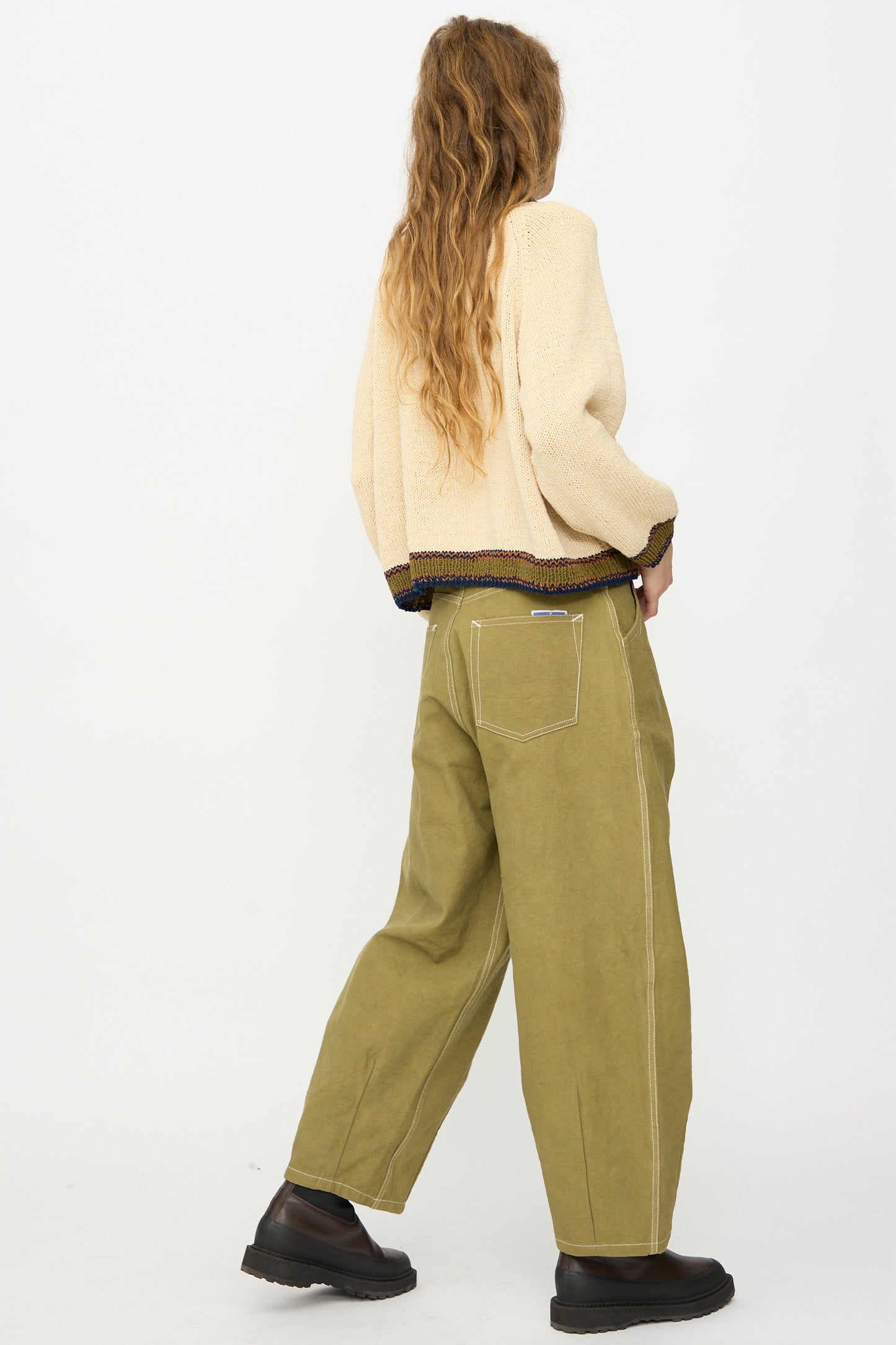 Person with long wavy hair wears a cream sweater and Story mfg. Canvas Lush Pant in Olive Wonky-Wear, featuring visible stitching, paired with black shoes, standing and facing away.