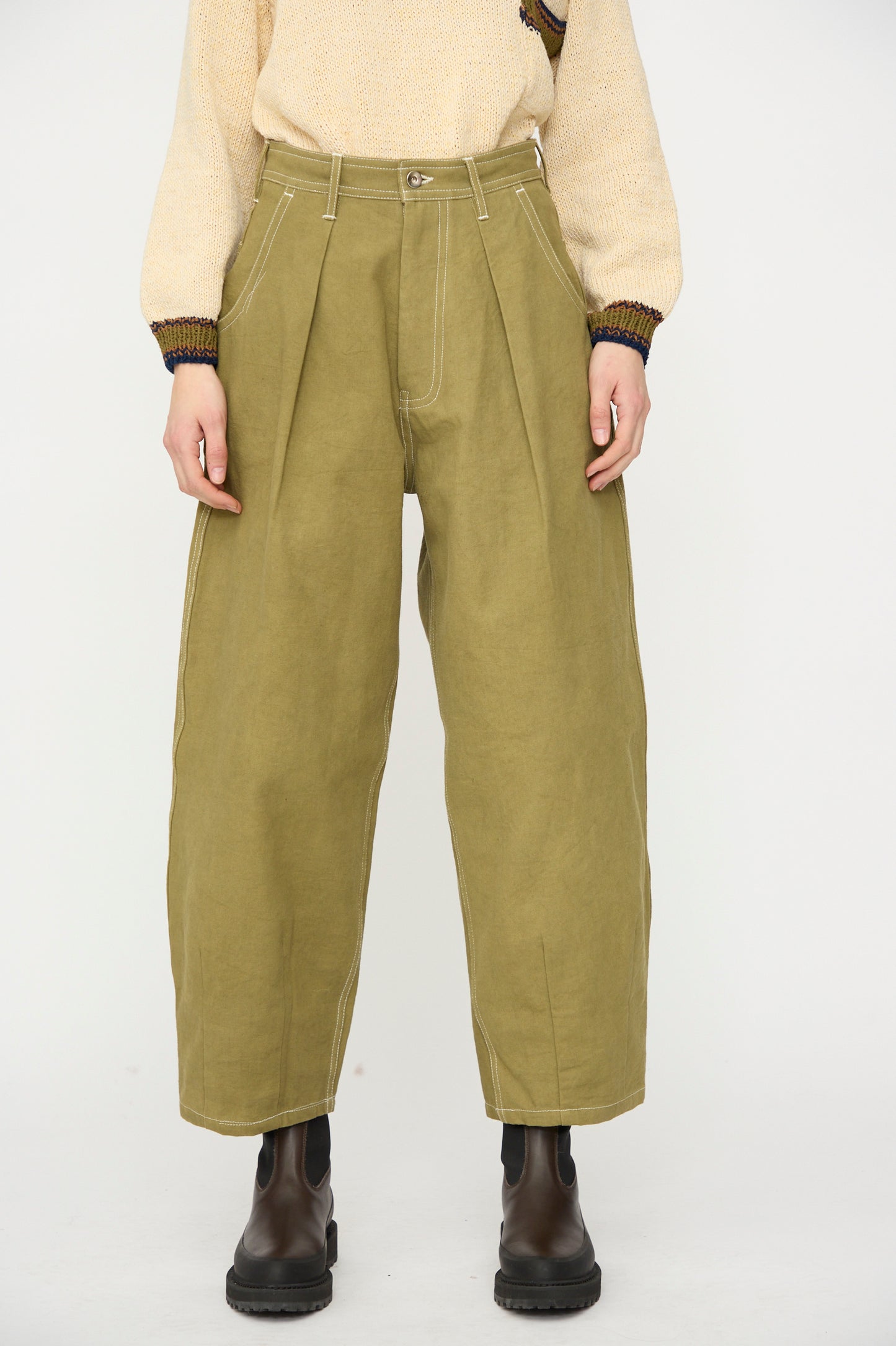 Person wearing Story mfg.'s Canvas Lush Pant in Olive Wonky-Wear, a beige knit sweater, and black ankle boots, standing against a plain white background.