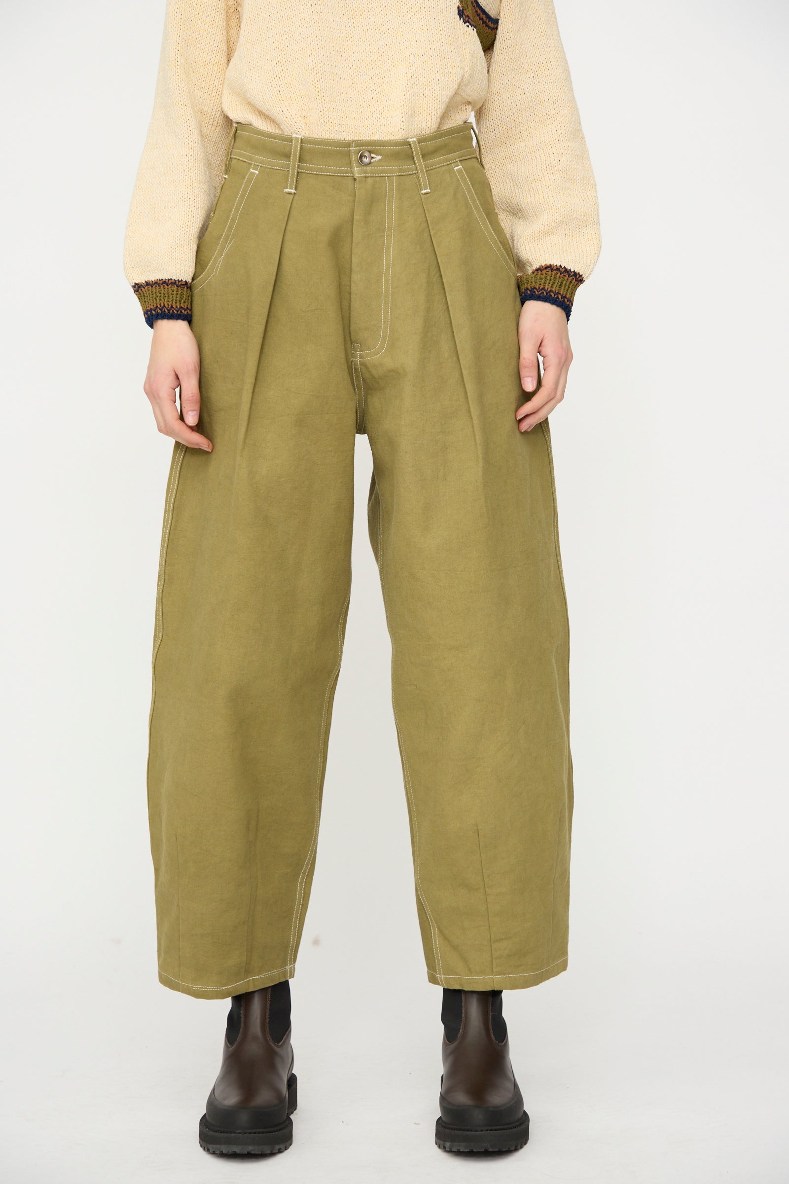 Person wearing Story mfg.'s Canvas Lush Pant in Olive Wonky-Wear, a beige knit sweater, and black ankle boots, standing against a plain white background.