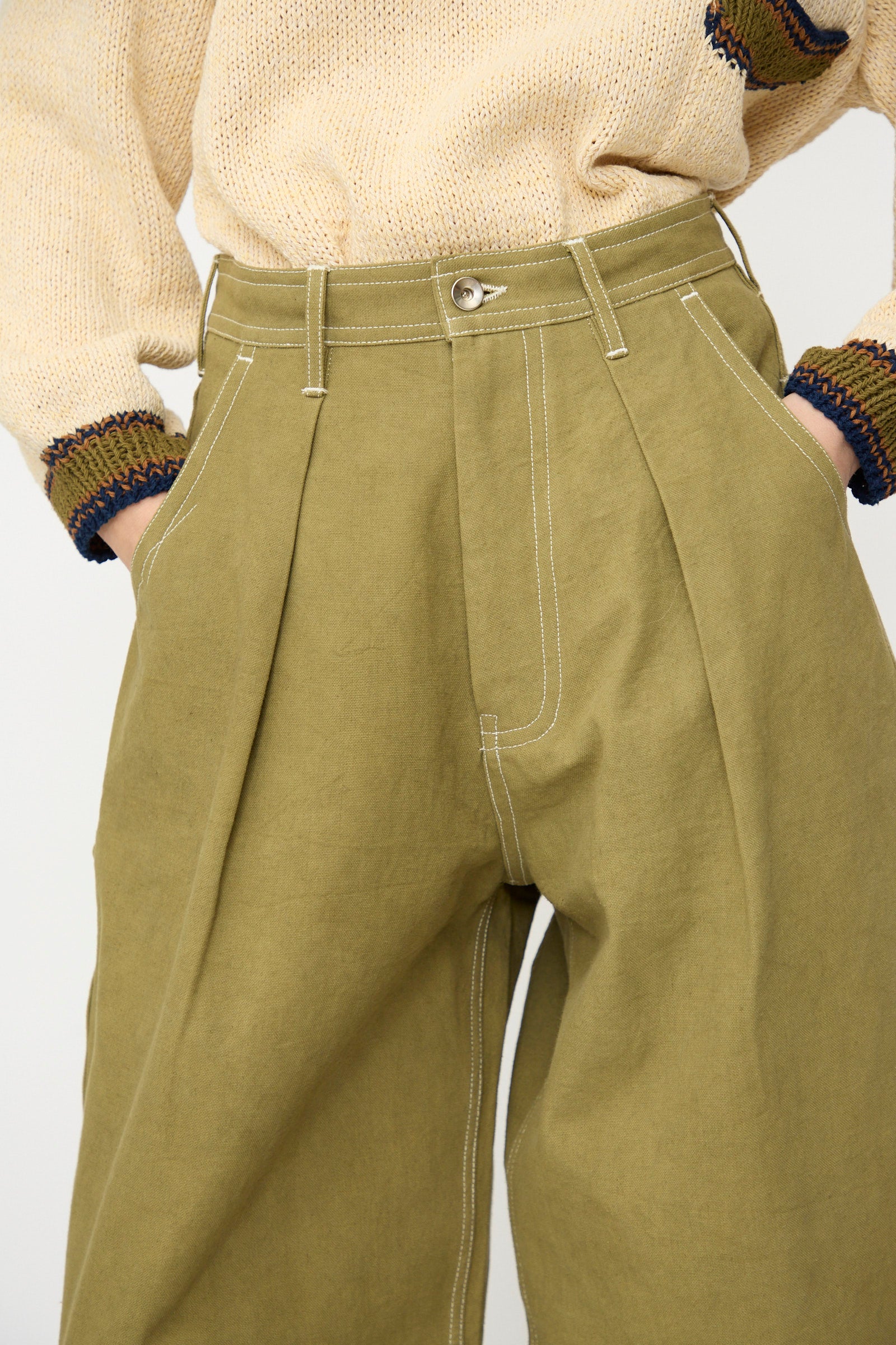 Person wearing Story mfg.'s Canvas Lush Pant in Olive Wonky-Wear, a relaxed fit, high-waisted, wide-leg workwear style with white stitching, paired with a cream knit sweater with multicolored cuffs and hands in pockets.