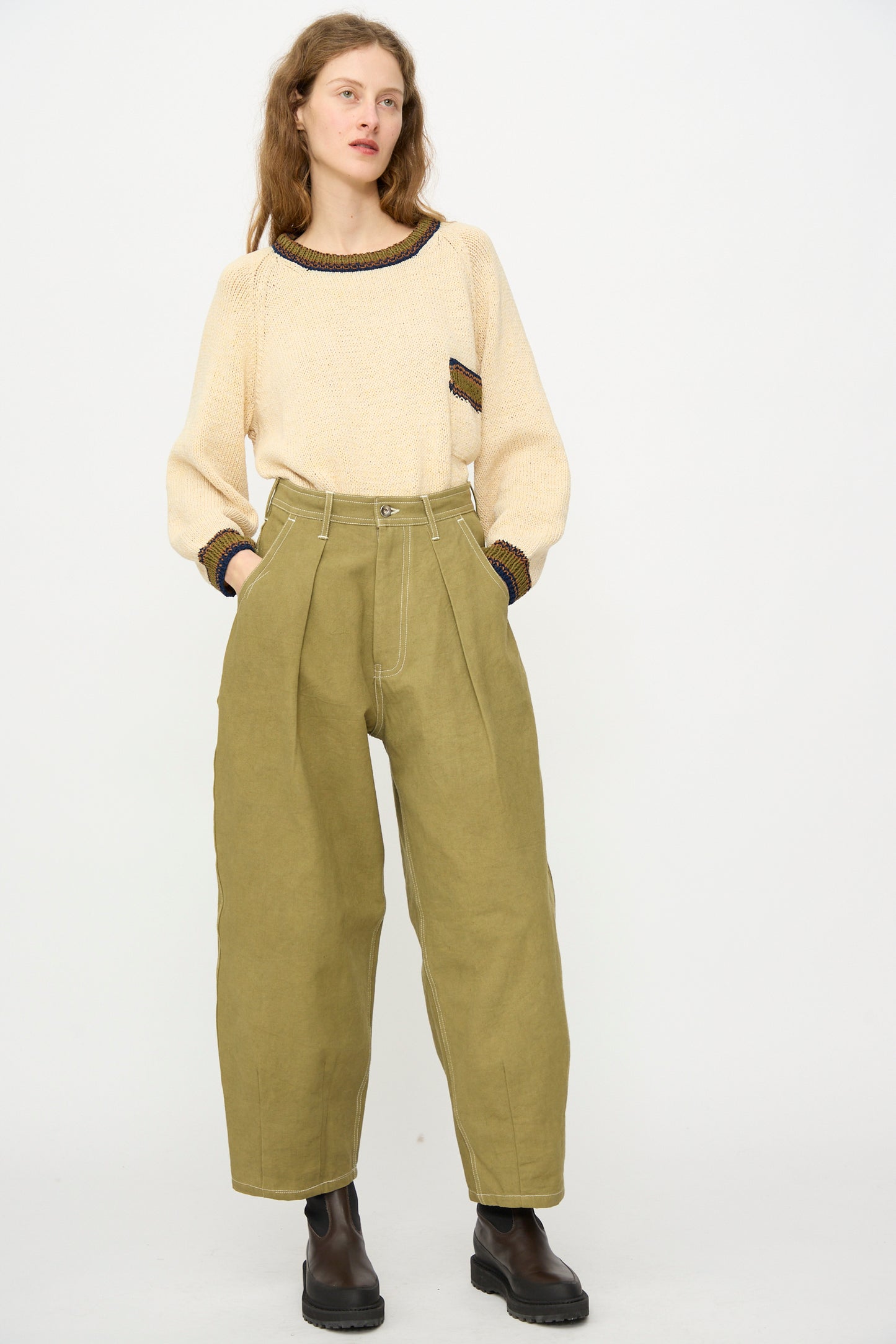 A person stands against a plain background wearing a cream sweater with dark trim, black boots, and the Story mfg. Canvas Lush Pant in Olive Wonky-Wear.