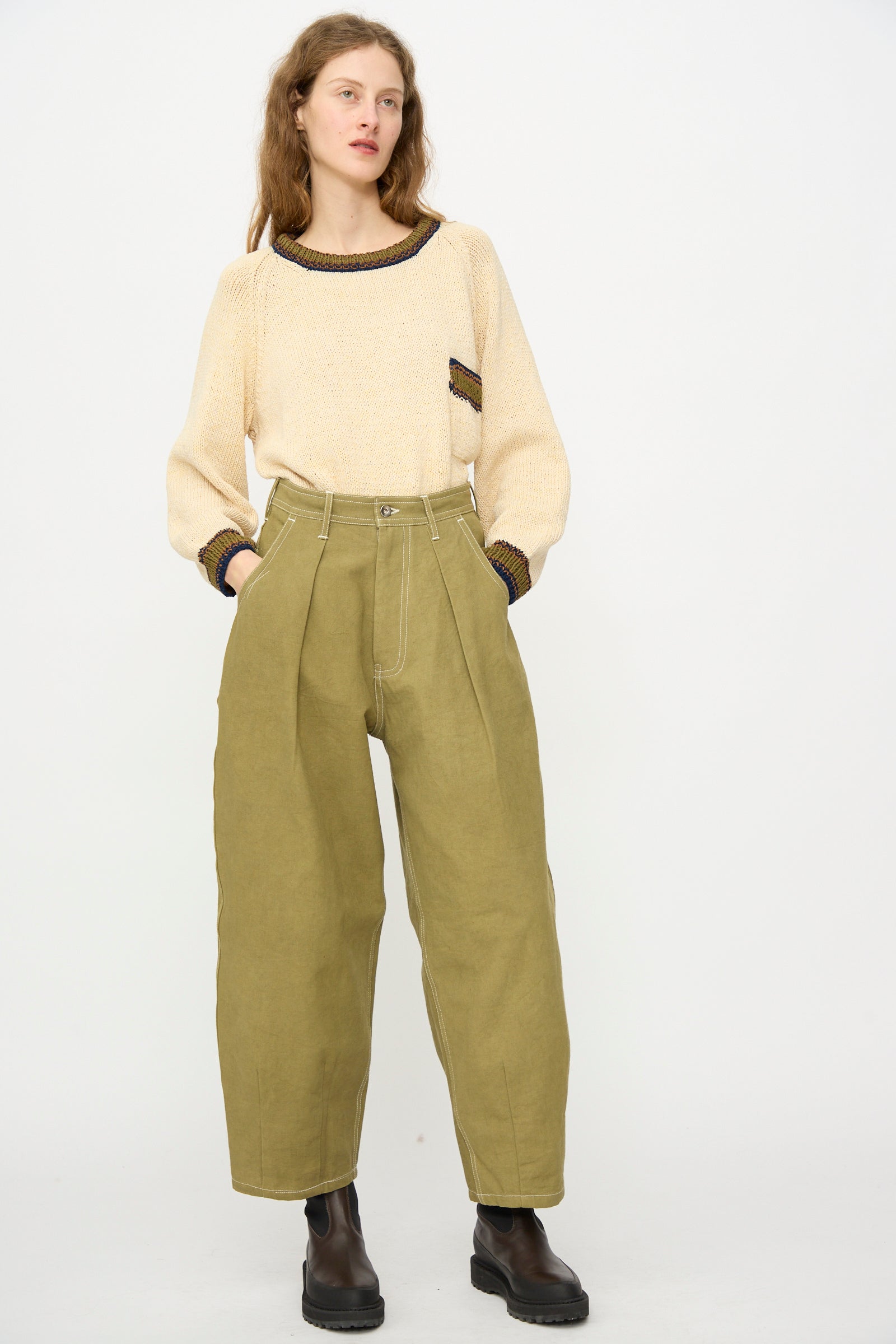 A person stands against a plain background wearing a cream sweater with dark trim, black boots, and the Story mfg. Canvas Lush Pant in Olive Wonky-Wear.