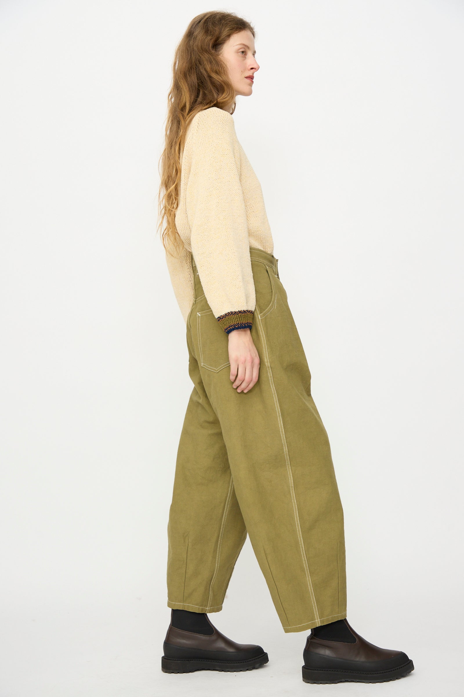 A person stands in profile wearing a beige sweater, Story mfg.'s Canvas Lush Pant in Olive Wonky-Wear with visible stitching, and black ankle boots against a plain white background.