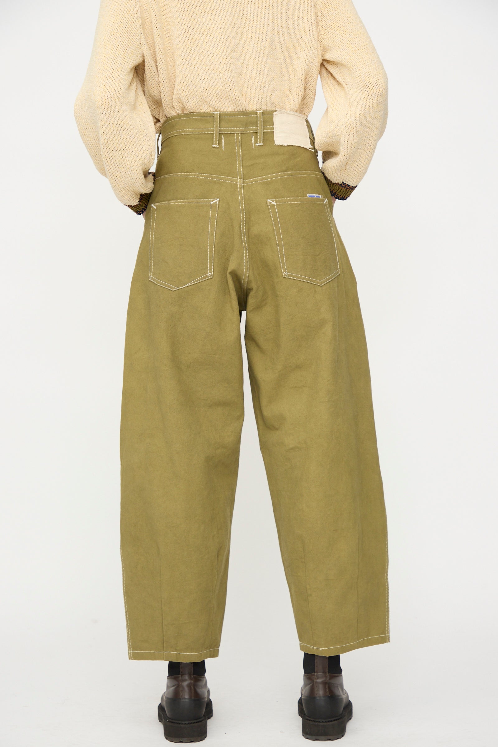 Person wearing Story mfg. Canvas Lush Pant in Olive Wonky-Wear with white stitching, black boots, and a beige sweater, shown from the back against a plain white background.
