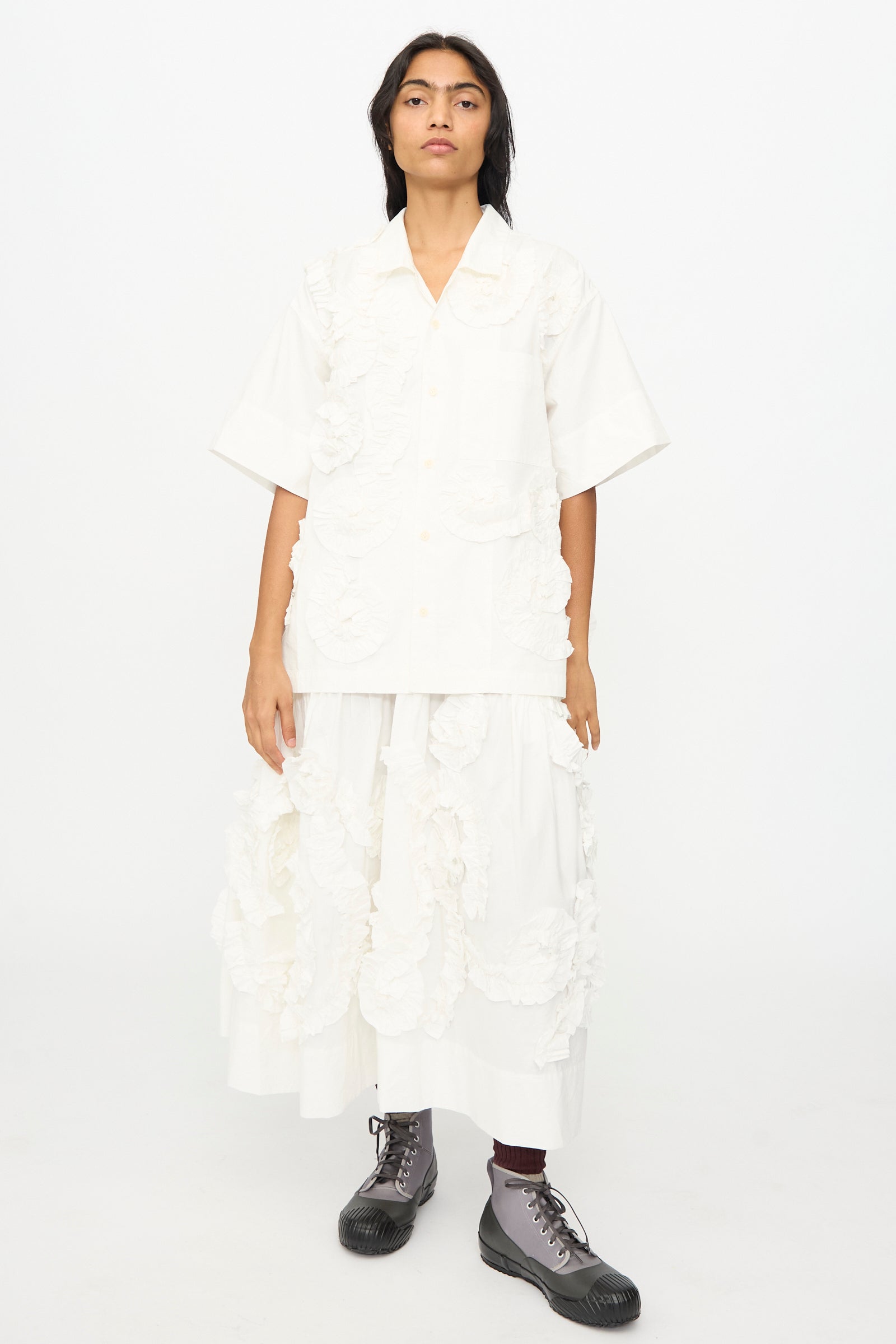 A person wears the oversized Story mfg. Hand Applique Greetings Shirt in Ecru Fossil with a textured floral skirt and gray lace-up boots, standing against a plain white background.