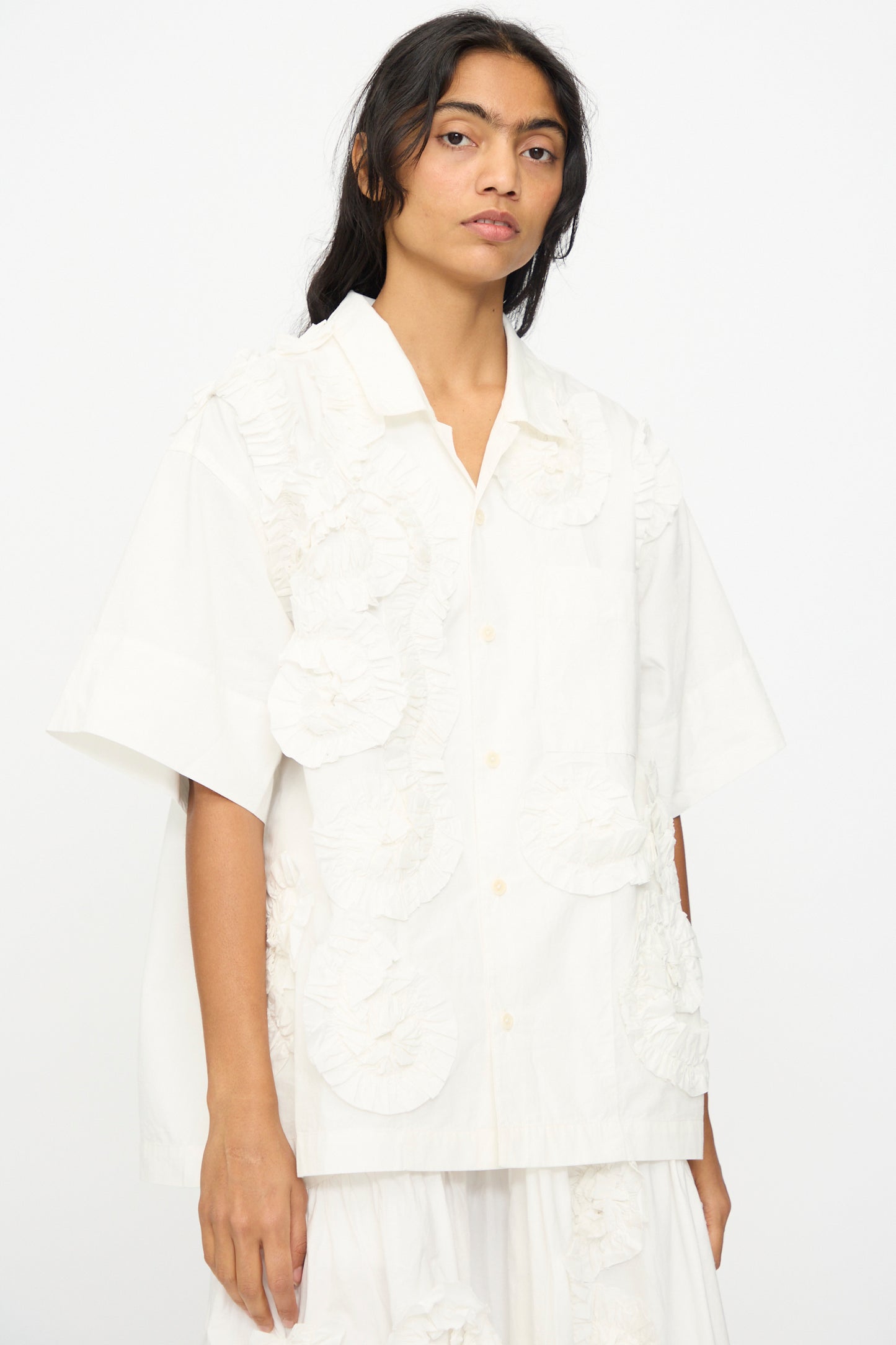 A person wears the Hand Applique Greetings Shirt in Ecru Fossil by Story mfg.—an oversized, short-sleeved white shirt with textured circular ruffle appliques—standing against a plain white background.