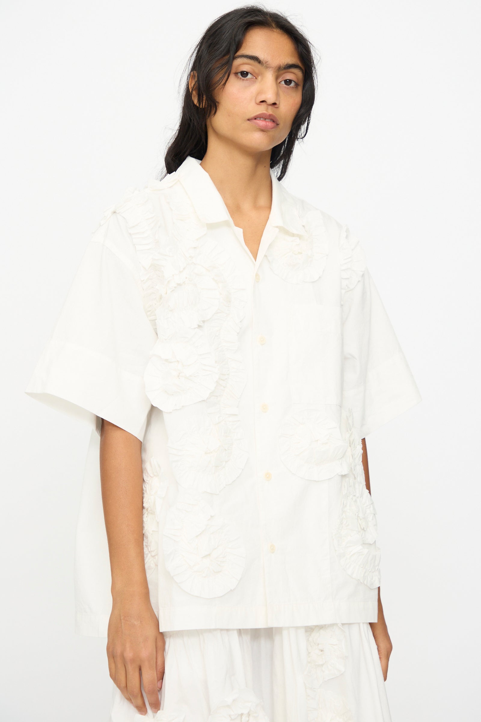 A person wears the Hand Applique Greetings Shirt in Ecru Fossil by Story mfg.—an oversized, short-sleeved white shirt with textured circular ruffle appliques—standing against a plain white background.