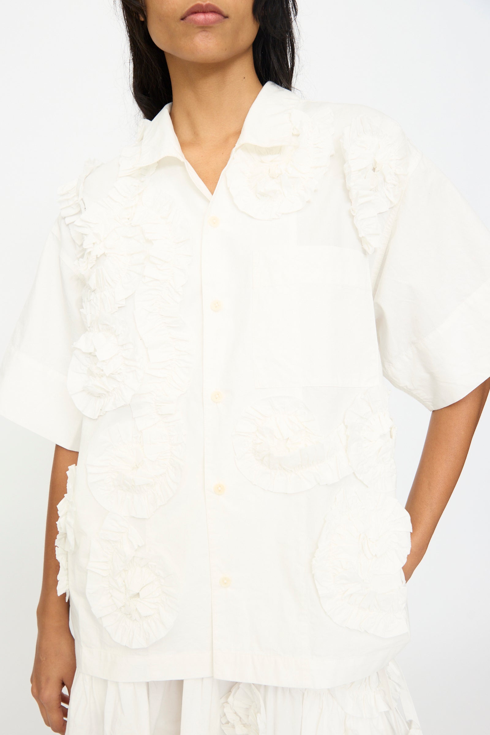 Someone wears the Story mfg. Hand Applique Greetings Shirt in Ecru Fossil, an oversized, short-sleeved piece featuring white textured ruffle applique details on the front and collar.