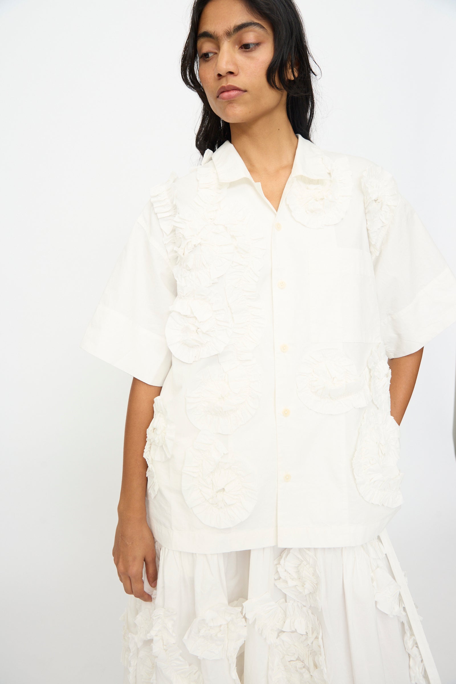 A woman wearing the Story mfg. Hand Applique Greetings Shirt in Ecru Fossil with textured ruffle details and a matching white skirt stands against a plain background.