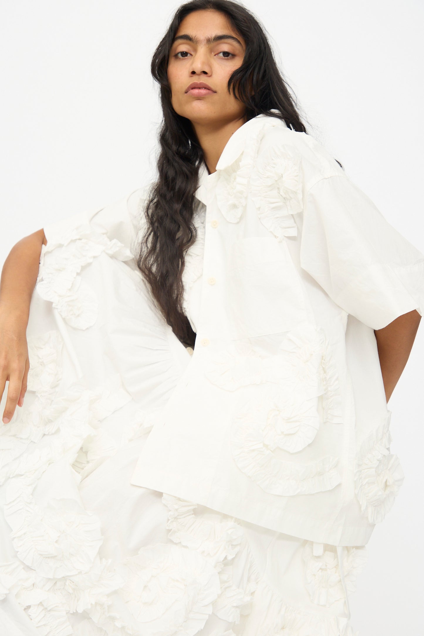 Wearing the Story mfg. Hand Applique Greetings Shirt in Ecru Fossil, an organic cotton white short-sleeve button-up with textured floral details, paired with a matching skirt, the model poses against a plain light background.