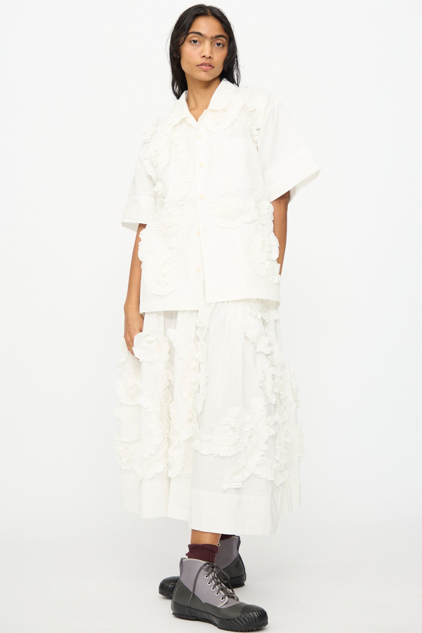 A person wearing a white short-sleeve button-up shirt and the Story mfg. Hand Applique Orbit Skirt in Ecru Fossil, paired with gray lace-up boots and brown socks, stands against a plain background.