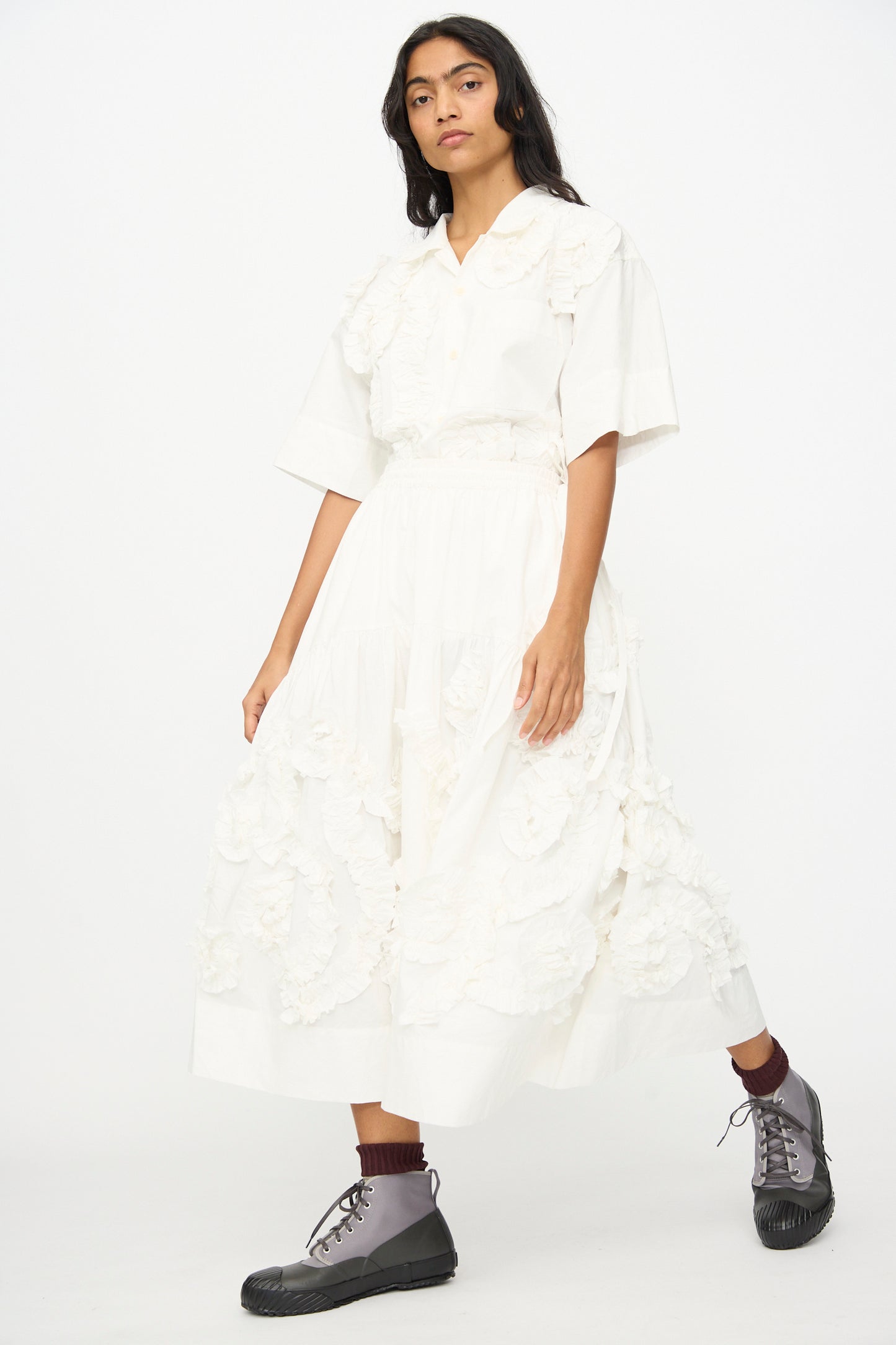 A person wears a white organic cotton ruffled short-sleeve blouse and the Story mfg. Hand Applique Orbit Skirt in Ecru Fossil, paired with brown socks and gray lace-up boots, standing against a plain white background.