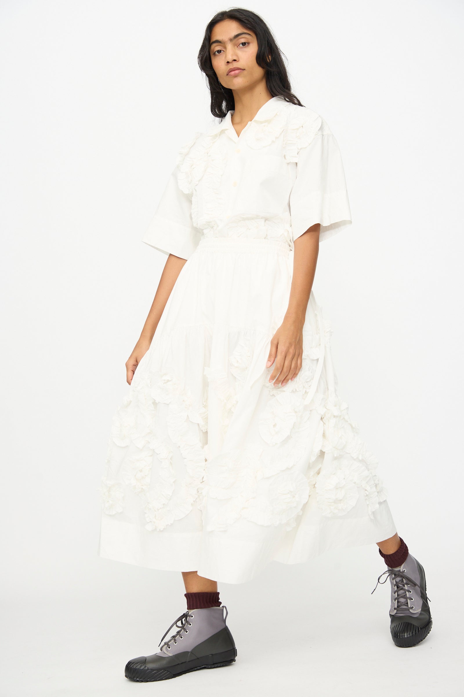 A person wears a white organic cotton ruffled short-sleeve blouse and the Story mfg. Hand Applique Orbit Skirt in Ecru Fossil, paired with brown socks and gray lace-up boots, standing against a plain white background.