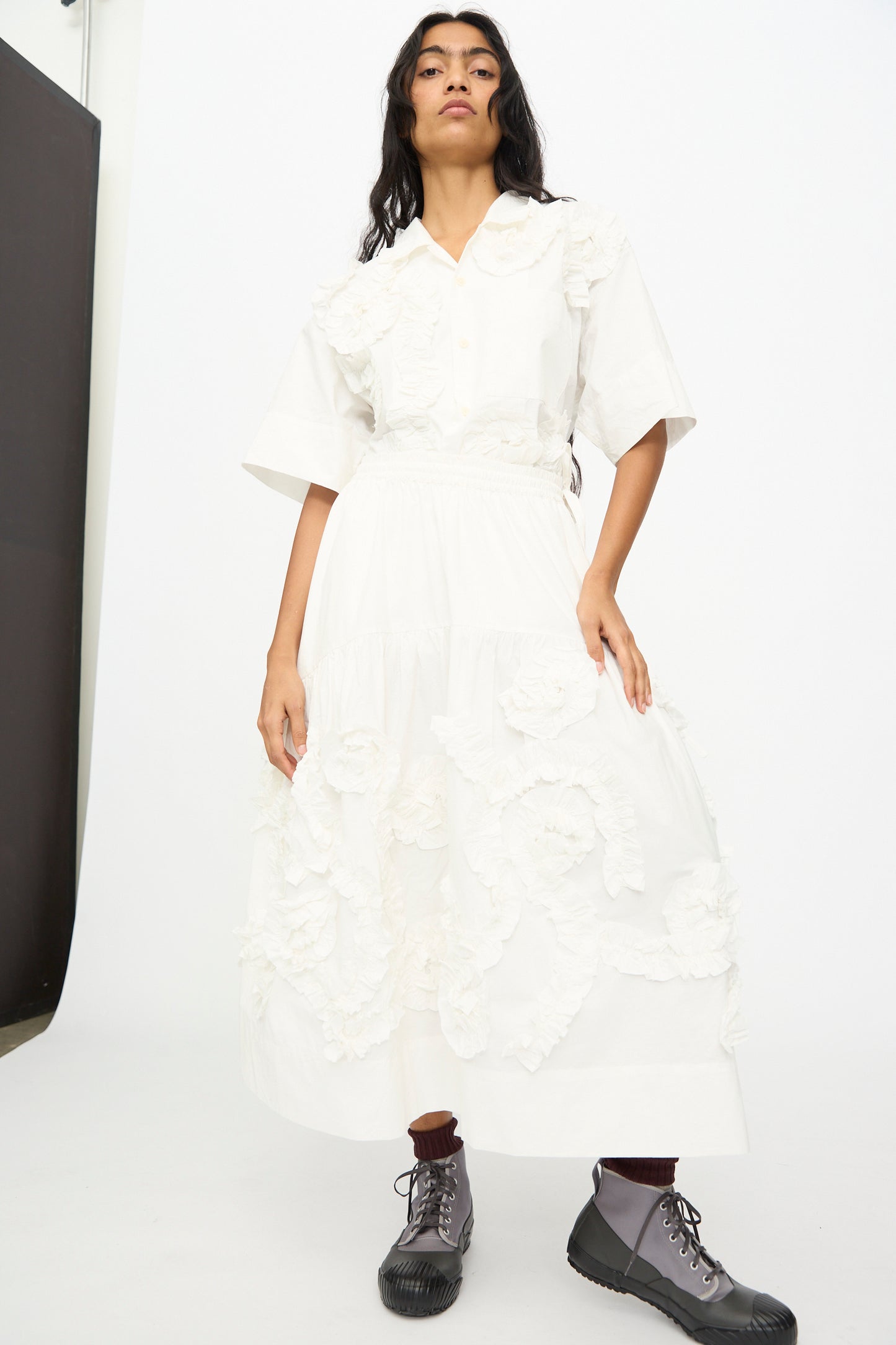 Someone stands against a white background wearing the Story mfg. Hand Applique Orbit Skirt in Ecru Fossil, featuring floral appliqué and subtle ruffle details, paired with gray lace-up boots.