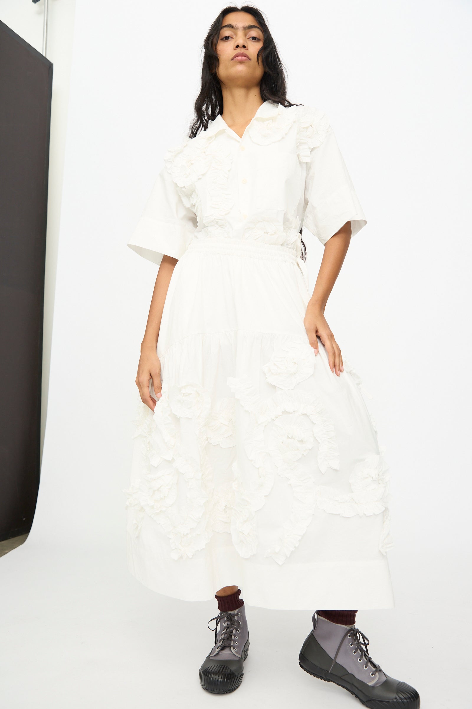 Someone stands against a white background wearing the Story mfg. Hand Applique Orbit Skirt in Ecru Fossil, featuring floral appliqué and subtle ruffle details, paired with gray lace-up boots.