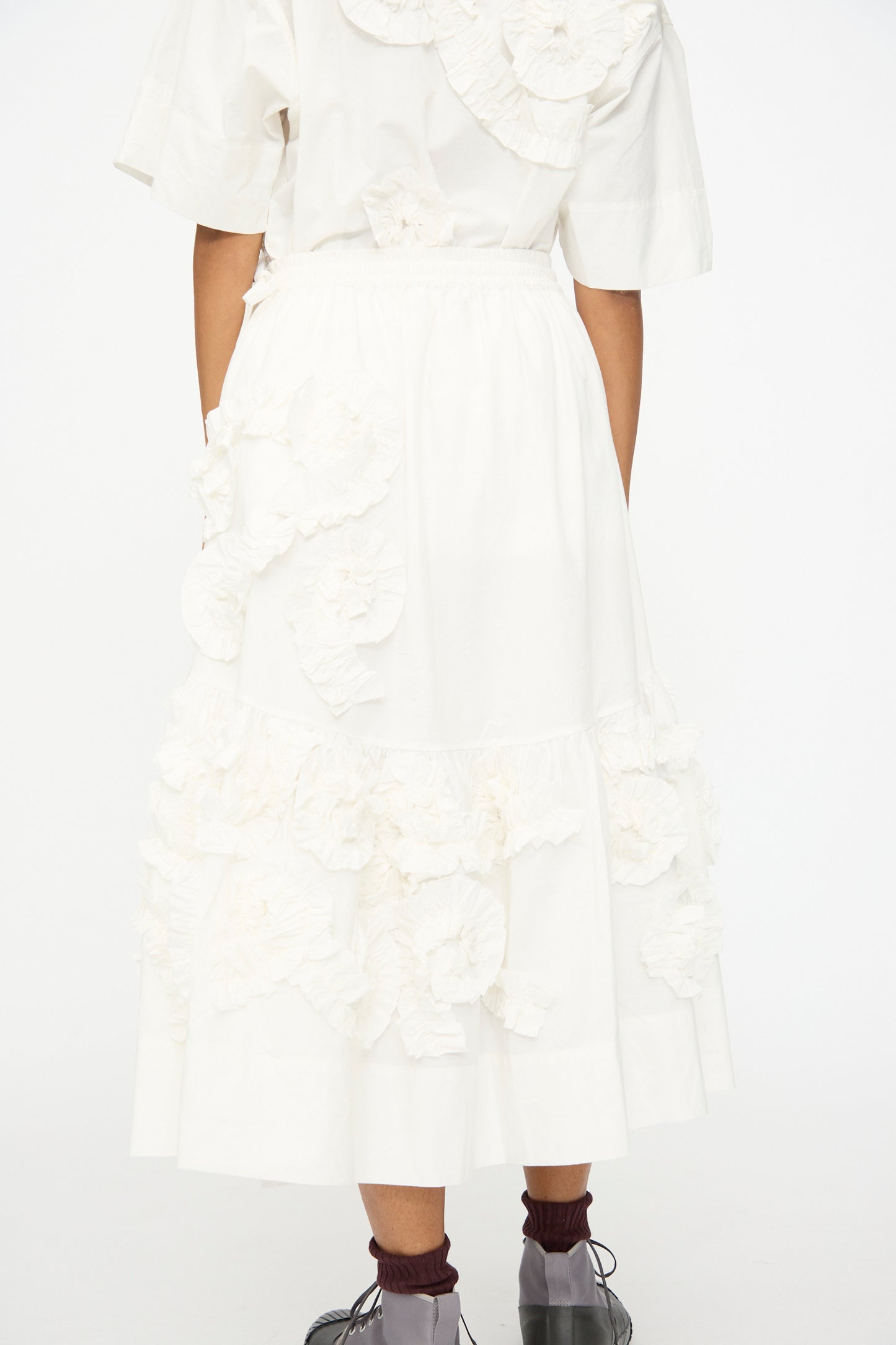 A person stands facing away in a white organic cotton top and the Story mfg. Hand Applique Orbit Skirt in Ecru Fossil, featuring ruffles and floral appliqués, paired with brown socks and black shoes against a plain background.