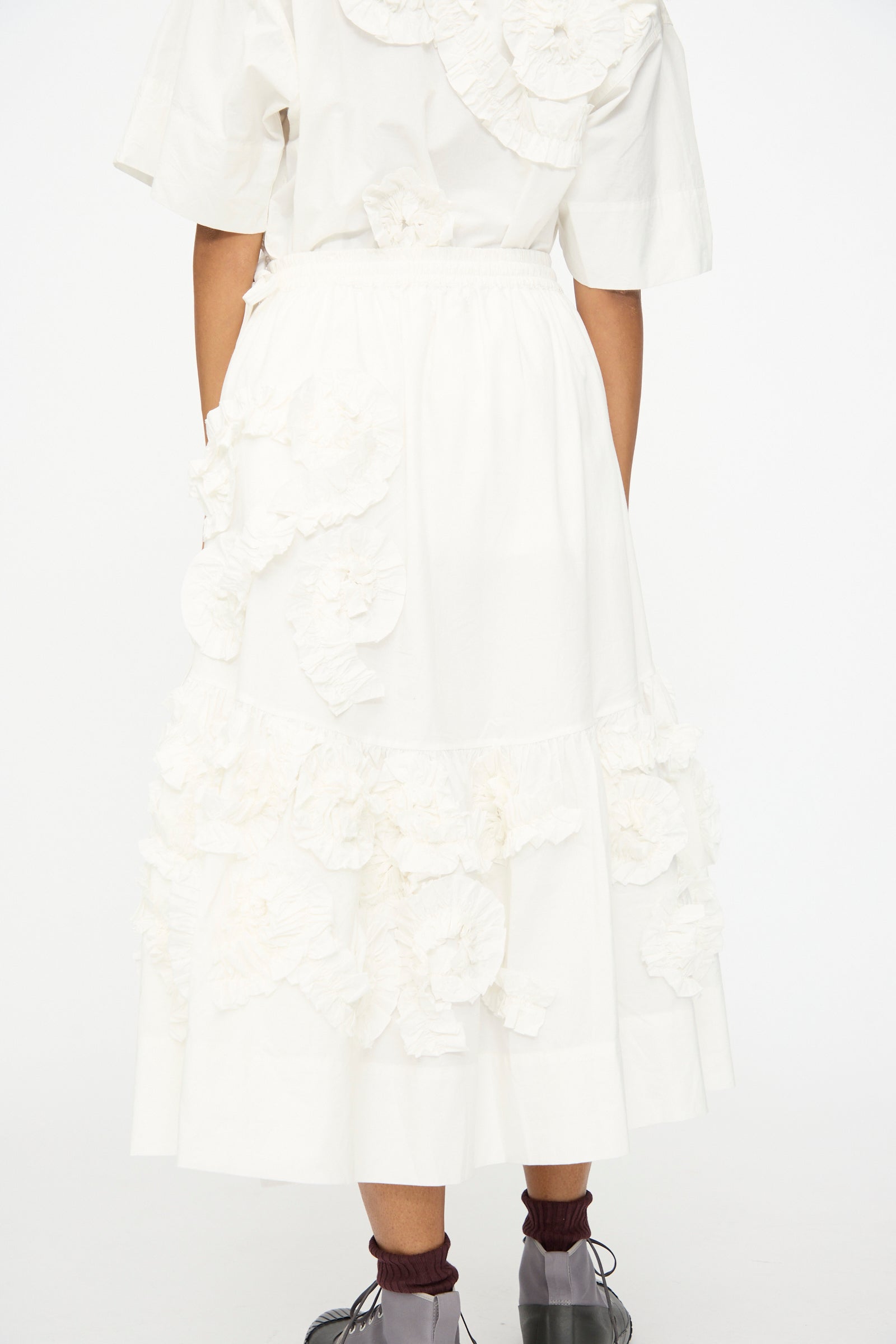 A person stands facing away in a white organic cotton top and the Story mfg. Hand Applique Orbit Skirt in Ecru Fossil, featuring ruffles and floral appliqués, paired with brown socks and black shoes against a plain background.