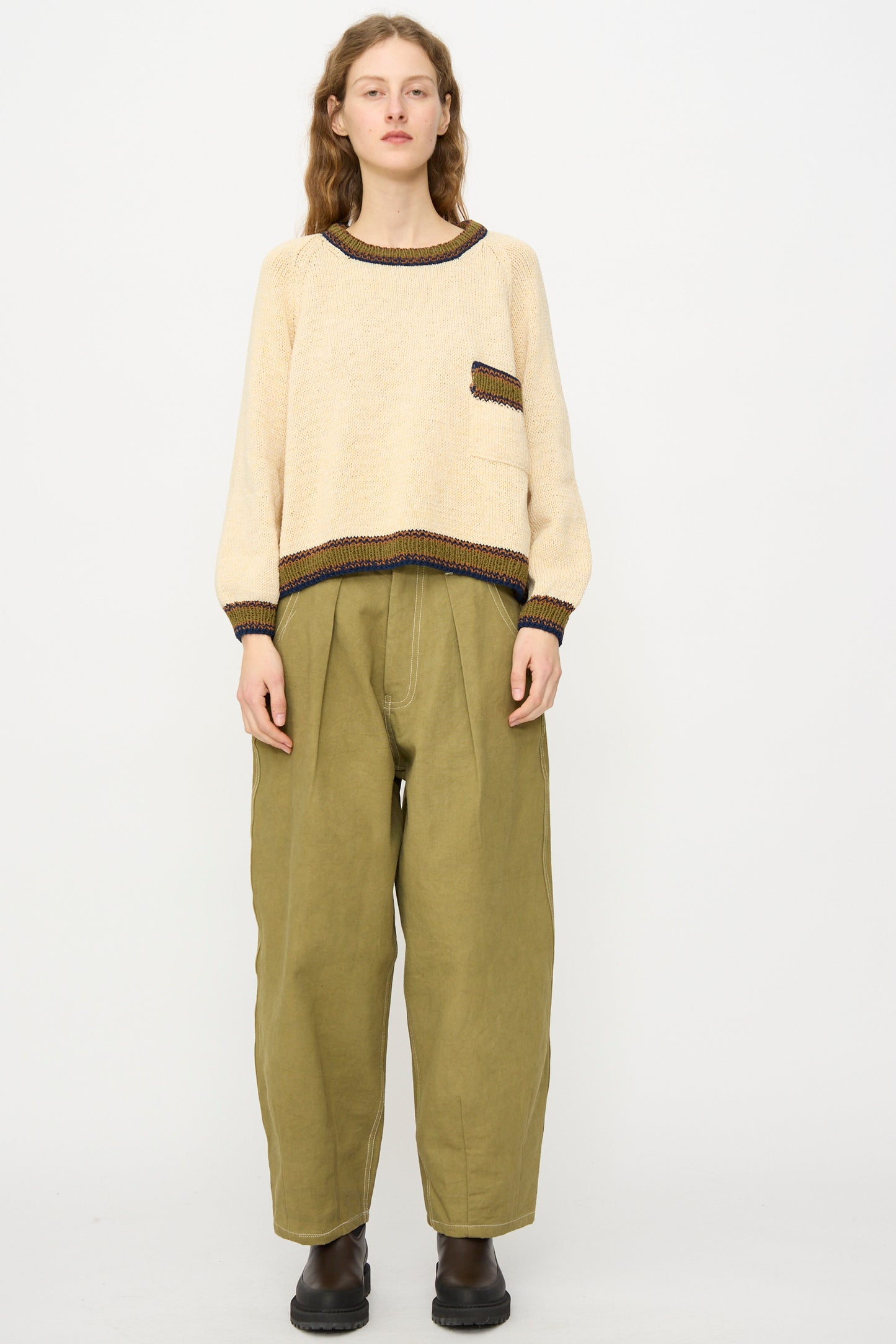 A person stands against a plain background wearing the Story mfg. Hand Knit Bulb Jumper in Twisted Ecru with olive green wide-leg pants and black shoes.