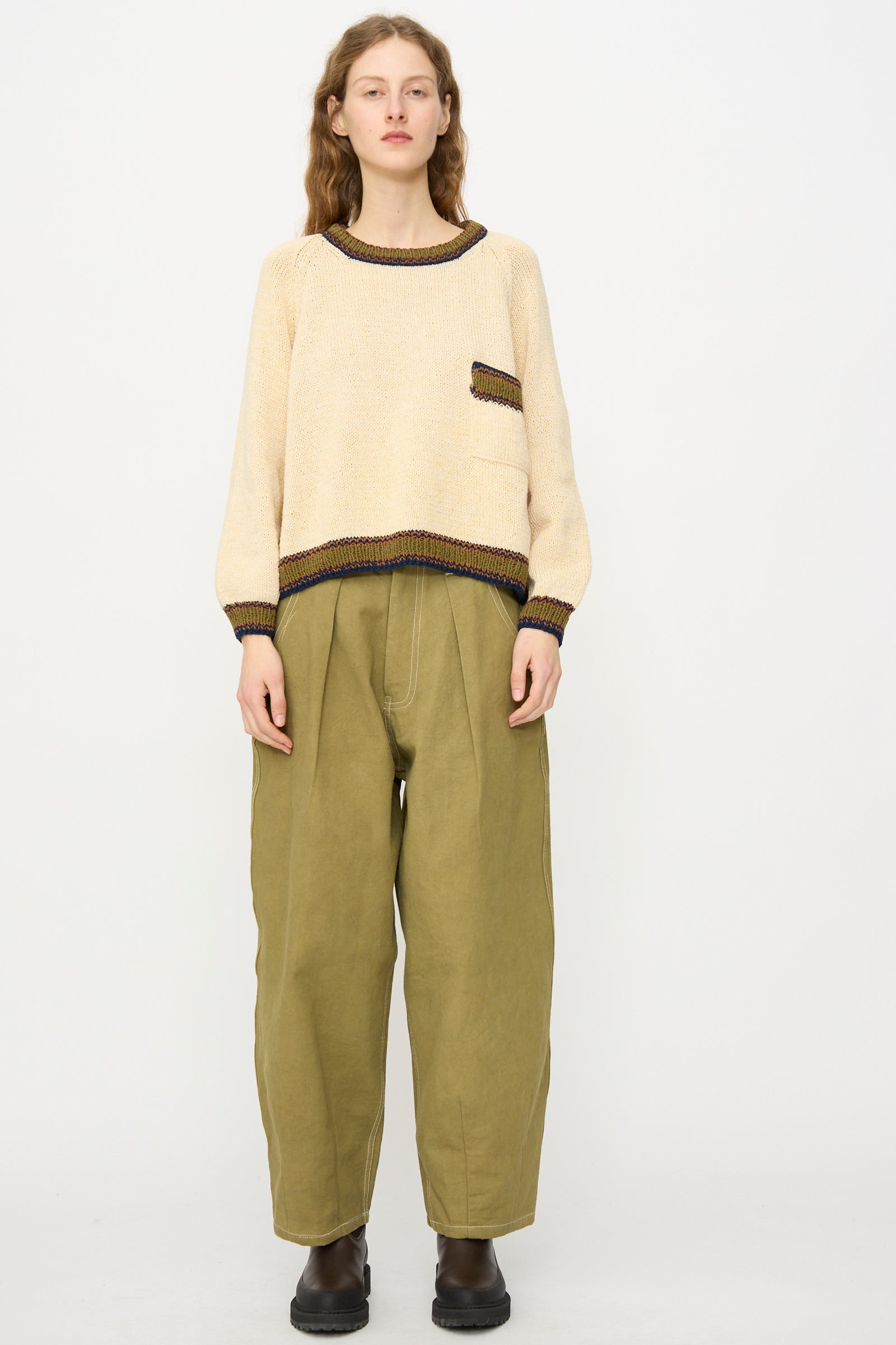 A person stands against a plain background wearing the Story mfg. Hand Knit Bulb Jumper in Twisted Ecru with olive green wide-leg pants and black shoes.