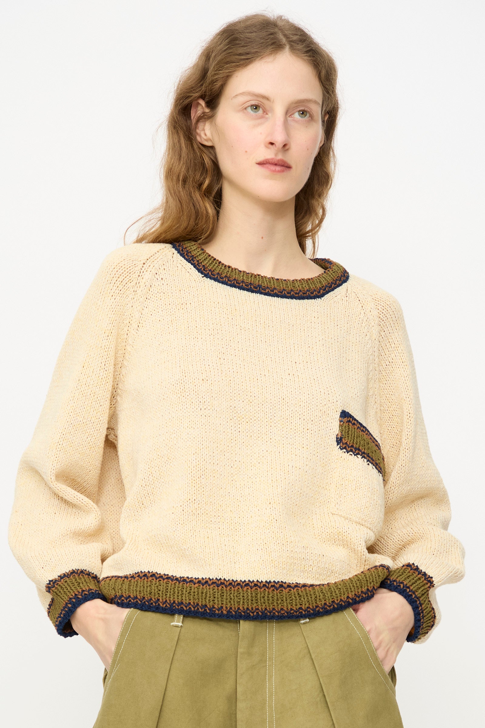 A person with long, wavy hair wears the Story mfg. Hand Knit Bulb Jumper in Twisted Ecru with contrasting trim and a small pocket, paired with olive green pleated pants, standing against a plain background.