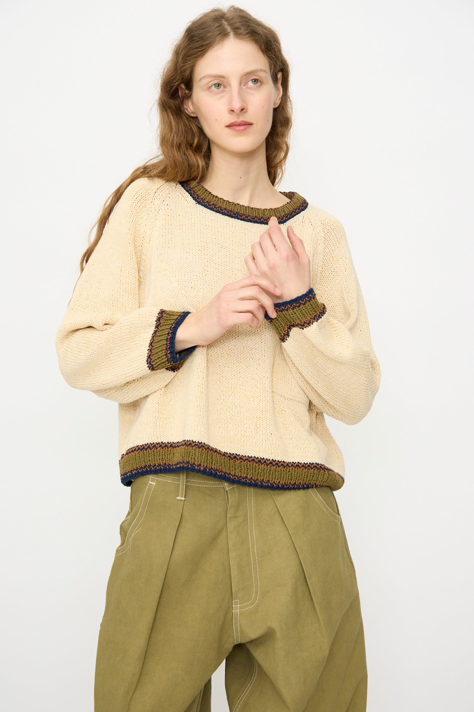 A person with long wavy hair models the Story mfg. Hand Knit Bulb Jumper in Twisted Ecru, paired with olive green pants, standing against a plain light backdrop.