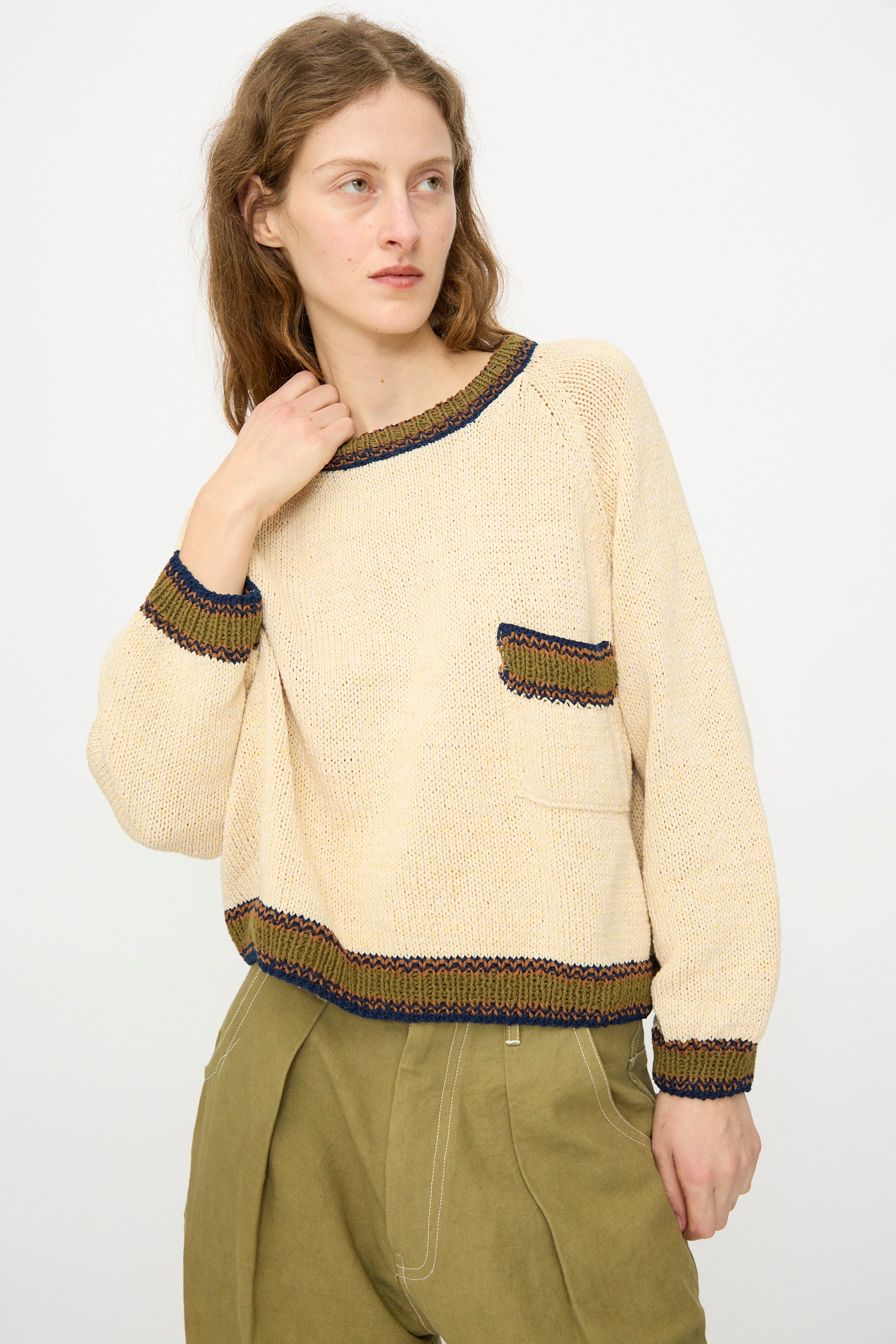 A woman wears the Story mfg. Hand Knit Bulb Jumper in Twisted Ecru, a cream cropped sweater with colored trim and a front pocket, paired with olive green pants, standing against a plain light background.