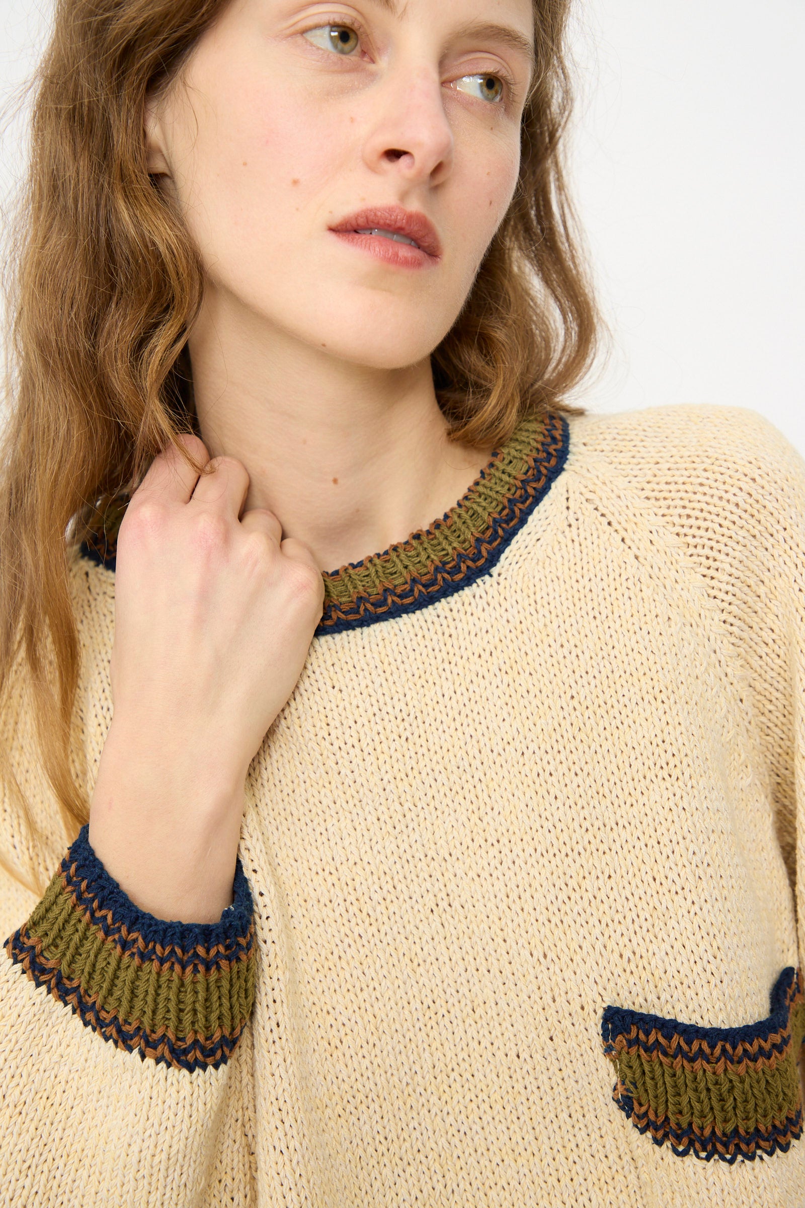 Woman wearing the Story mfg. Hand Knit Bulb Jumper in Twisted Ecru, featuring green and blue trim on the collar, cuffs, and pocket, looking slightly to the side.