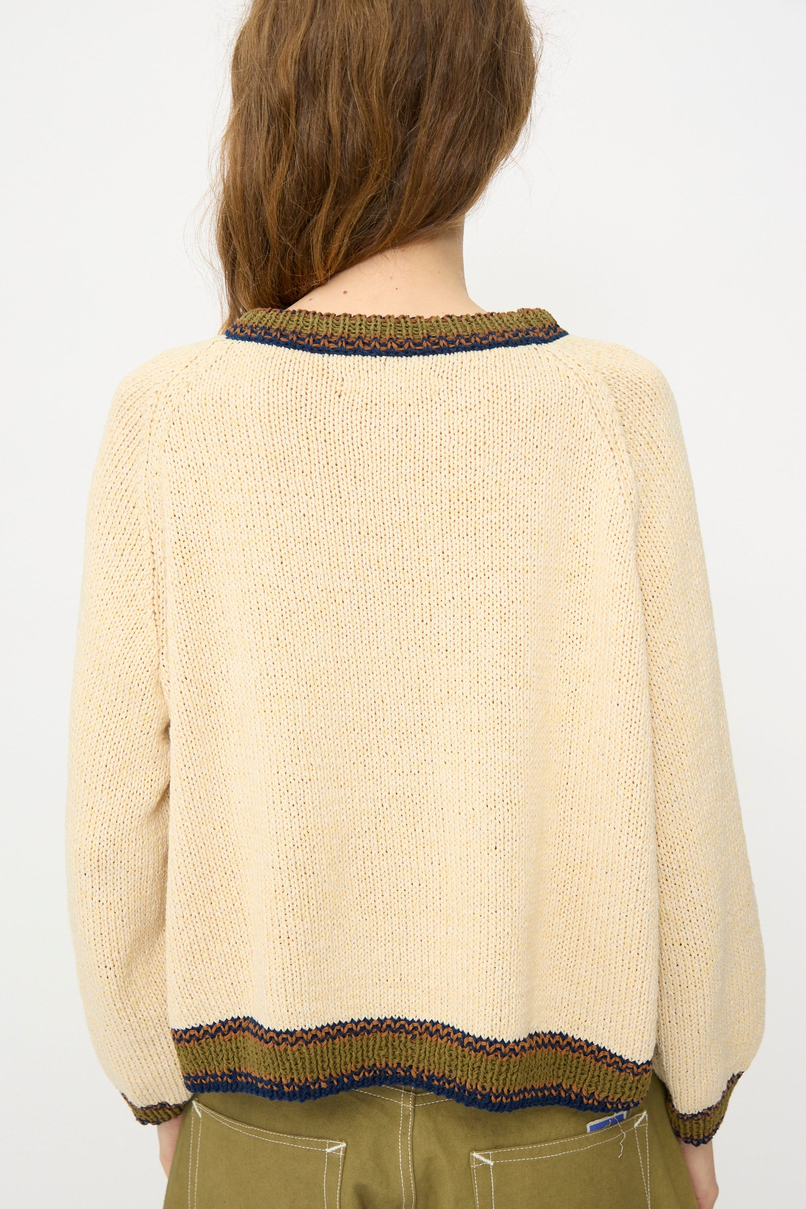 A person with long brown hair is seen from behind, wearing the Story mfg. Hand Knit Bulb Jumper in Twisted Ecru, which features green and brown trim, paired with olive green pants.