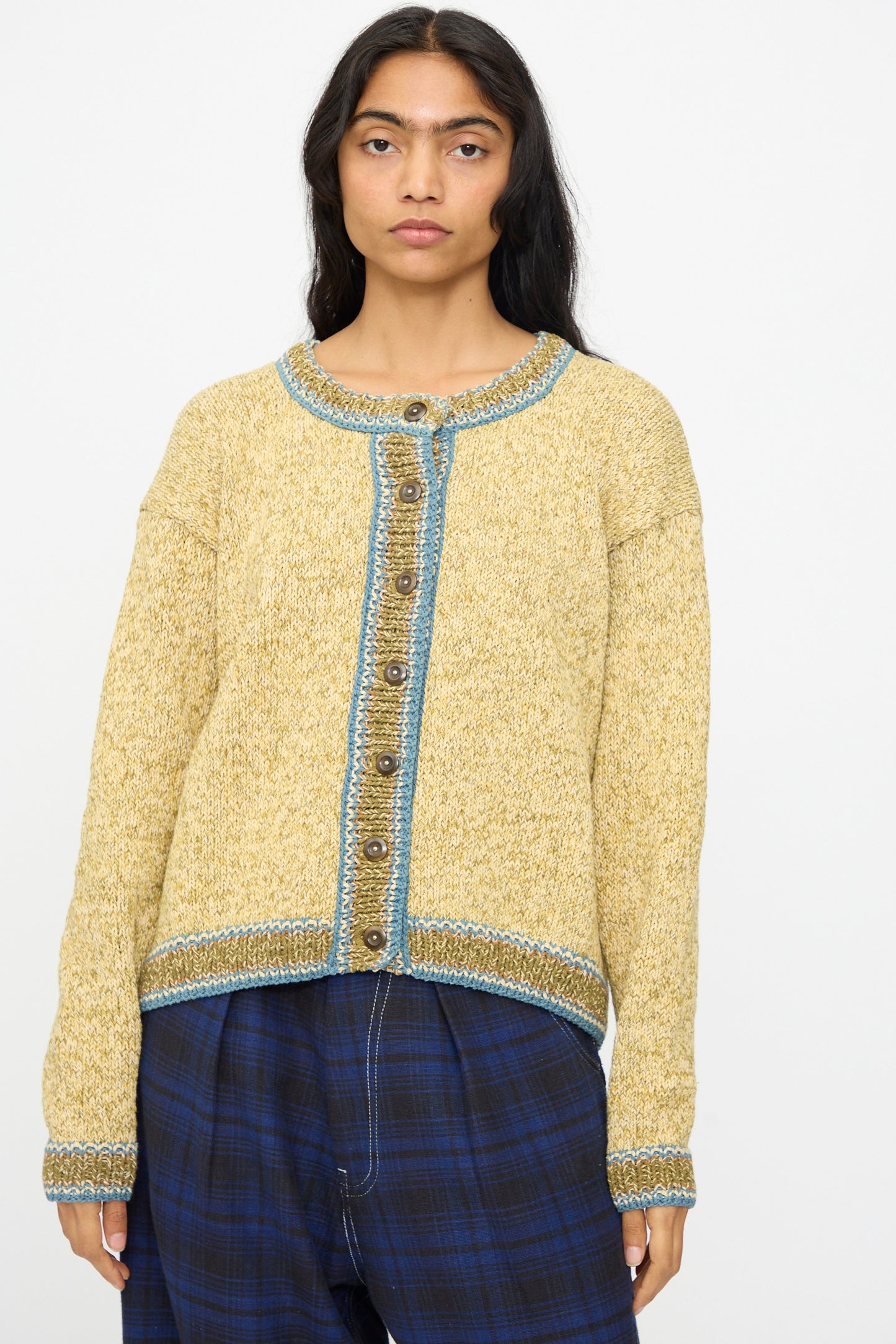 A person wears the Hand Knit Root Cardigan in Kelp by Story mfg.—a relaxed fit, handknit cotton cardigan in yellow with blue trim and buttons—paired with blue plaid pants against a plain white background.