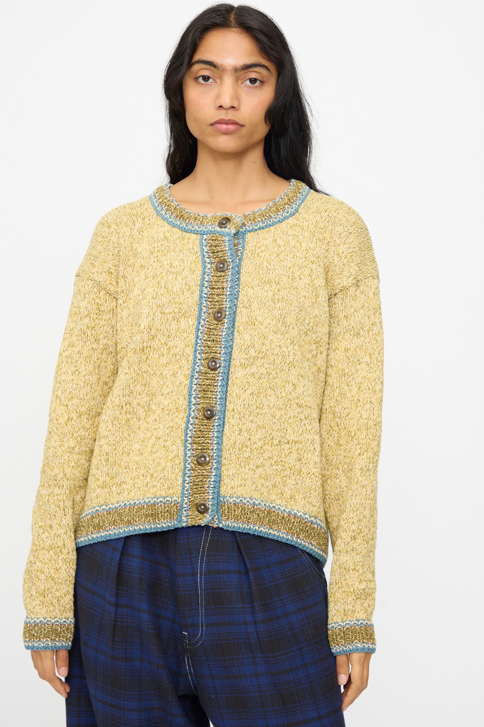 A person wears the Hand Knit Root Cardigan in Kelp by Story mfg.—a relaxed fit, handknit cotton cardigan in yellow with blue trim and buttons—paired with blue plaid pants against a plain white background.