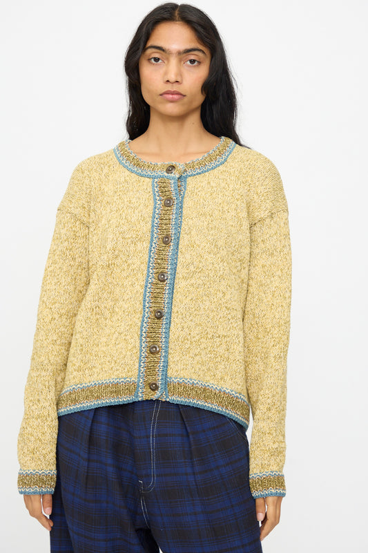 A person wears the Hand Knit Root Cardigan in Kelp by Story mfg.—a relaxed fit, handknit cotton cardigan in yellow with blue trim and buttons—paired with blue plaid pants against a plain white background.