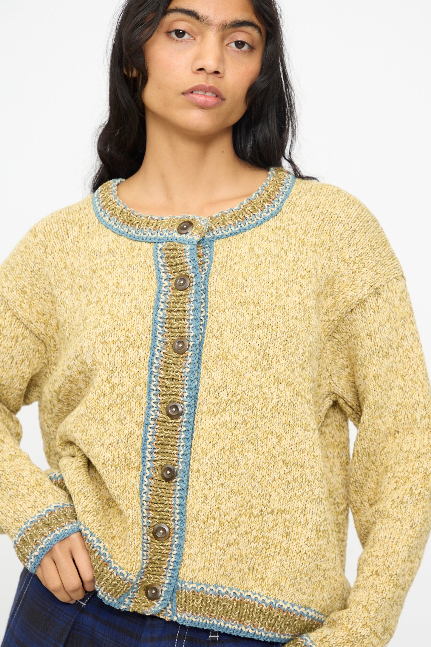 A woman wears the Story mfg. Hand Knit Root Cardigan in Kelp, a relaxed fit yellow cotton piece with blue trim and brown buttons, standing against a plain background.
