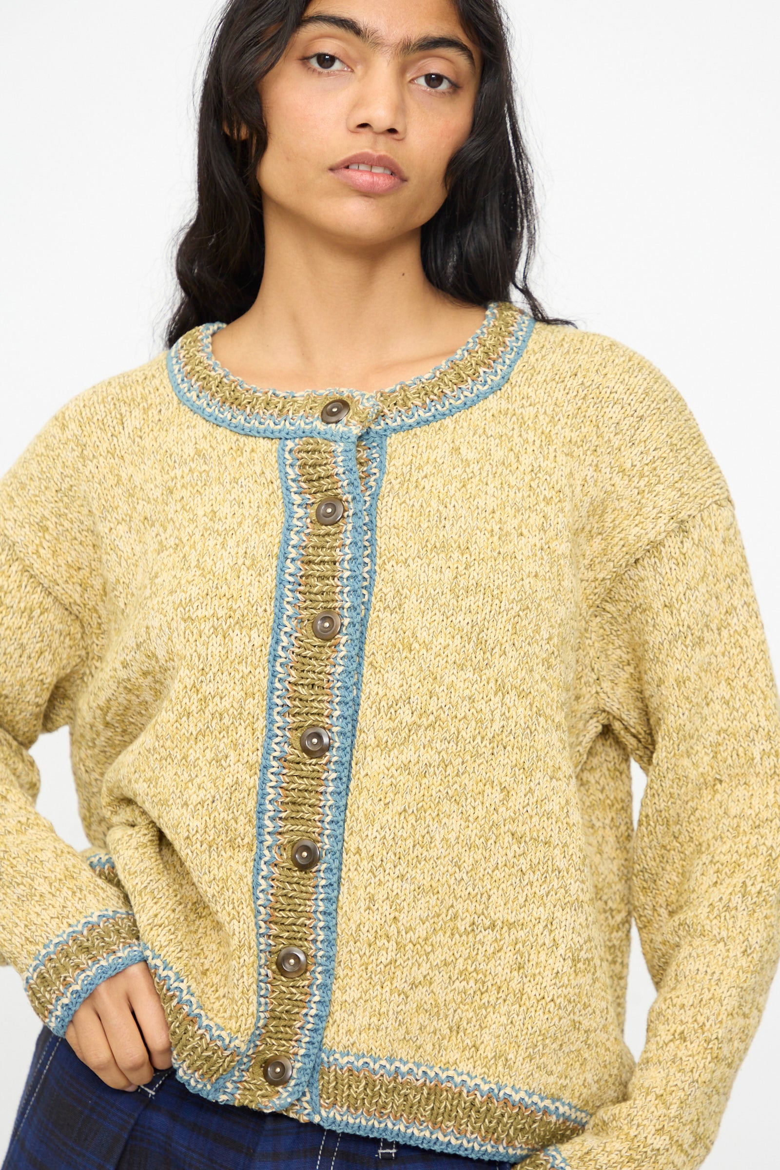 A woman wears the Story mfg. Hand Knit Root Cardigan in Kelp, a relaxed fit yellow cotton piece with blue trim and brown buttons, standing against a plain background.