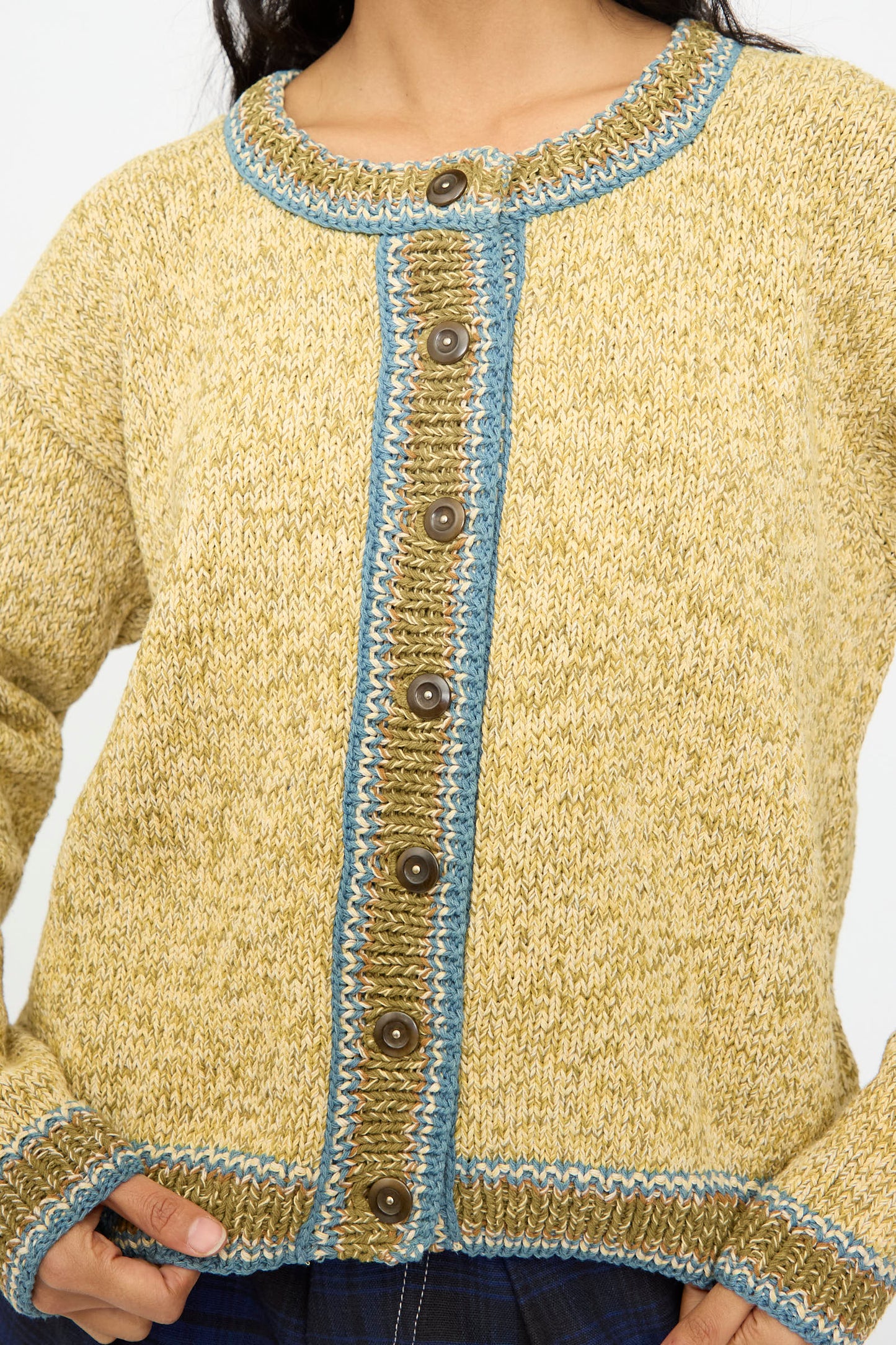 A person wears the Story mfg. Hand Knit Root Cardigan in Kelp, a relaxed fit yellow cardigan with brown buttons and blue trim, shown from neck to waist.