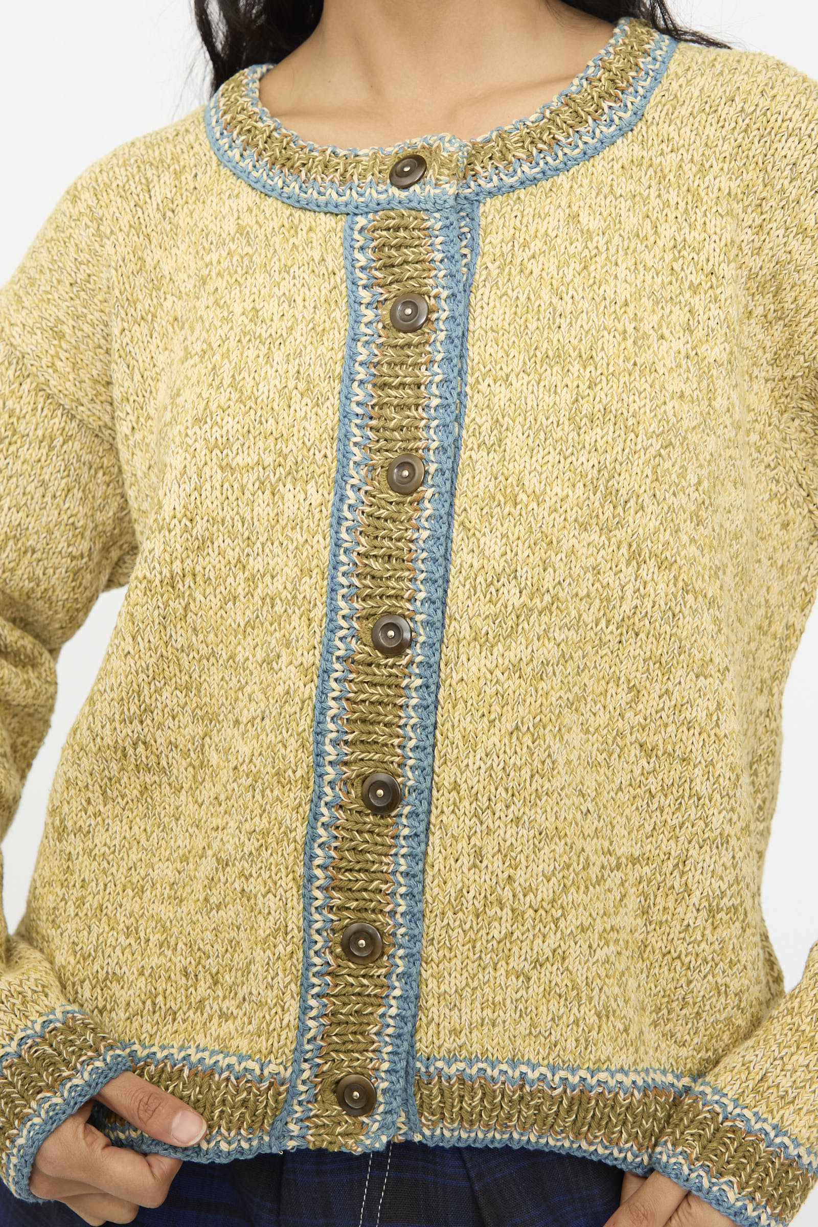 A person wears the Story mfg. Hand Knit Root Cardigan in Kelp, a relaxed fit yellow cardigan with brown buttons and blue trim, shown from neck to waist.