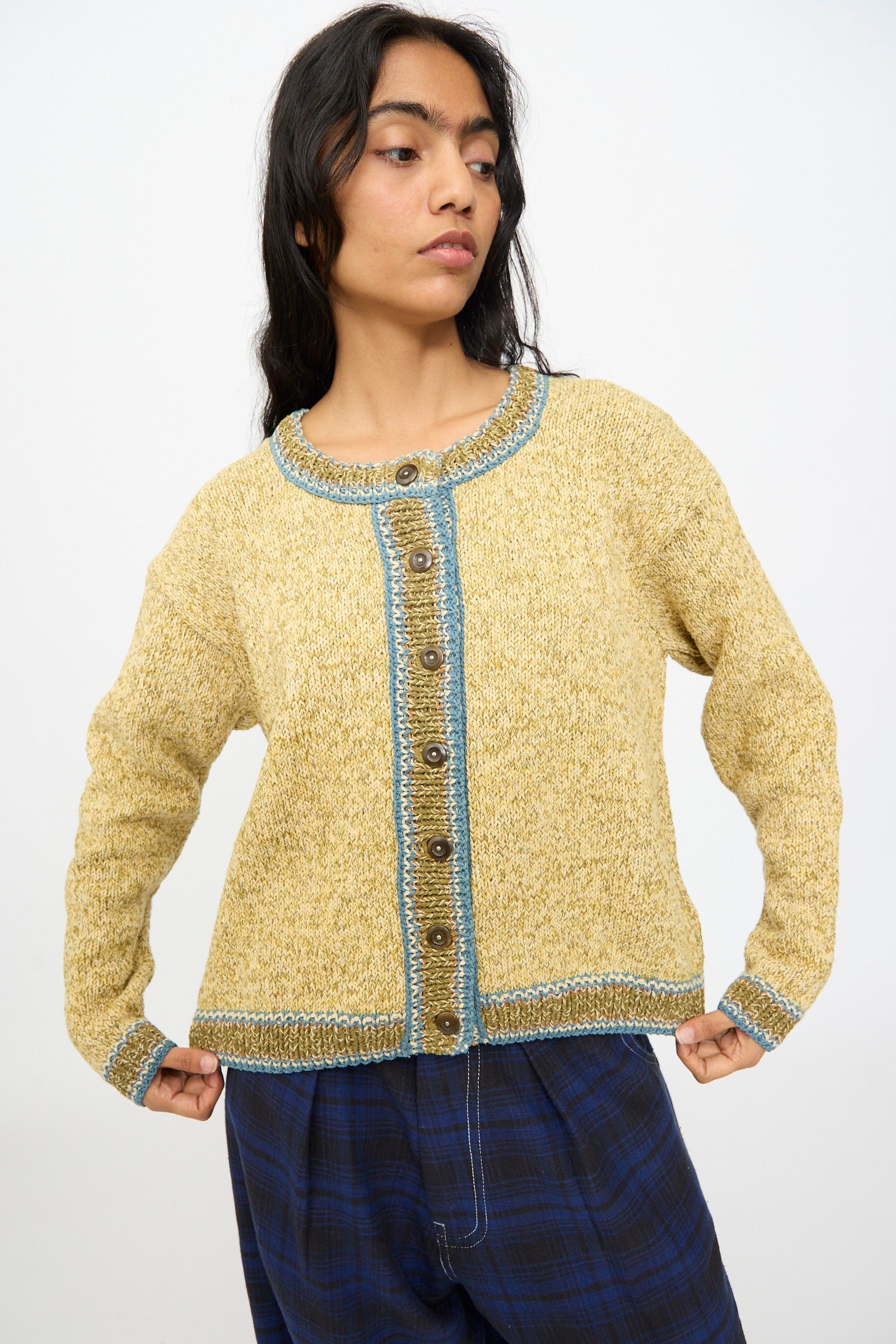 A person wears the Story mfg. Hand Knit Root Cardigan in Kelp—a relaxed fit yellow cotton cardigan with blue trim and brown buttons—paired with blue plaid pants, standing against a plain light background.