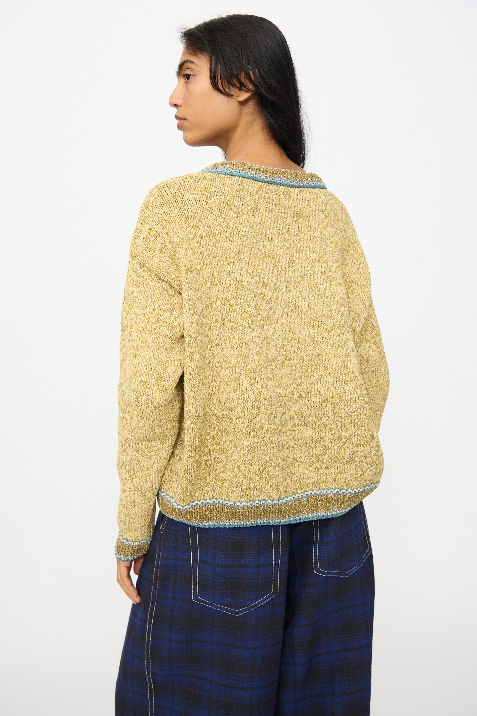 Person wearing the Hand Knit Root Cardigan in Kelp by Story mfg., featuring loose-knit yellow with blue trim, paired with blue plaid pants and standing with their back to the camera against a plain background.