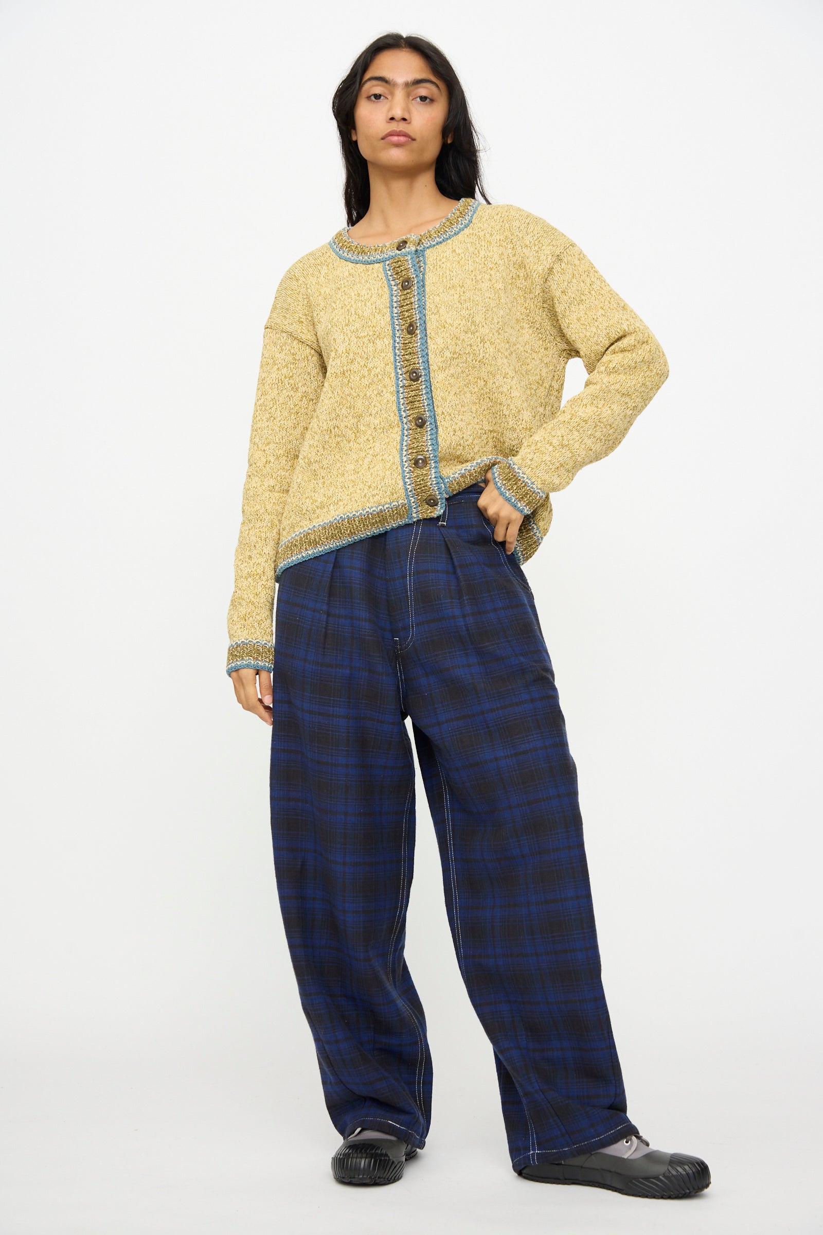 A person wears the Story mfg. Hand Knit Root Cardigan in Kelp, styled with blue plaid wide-leg pants and black shoes, standing against a plain white background.
