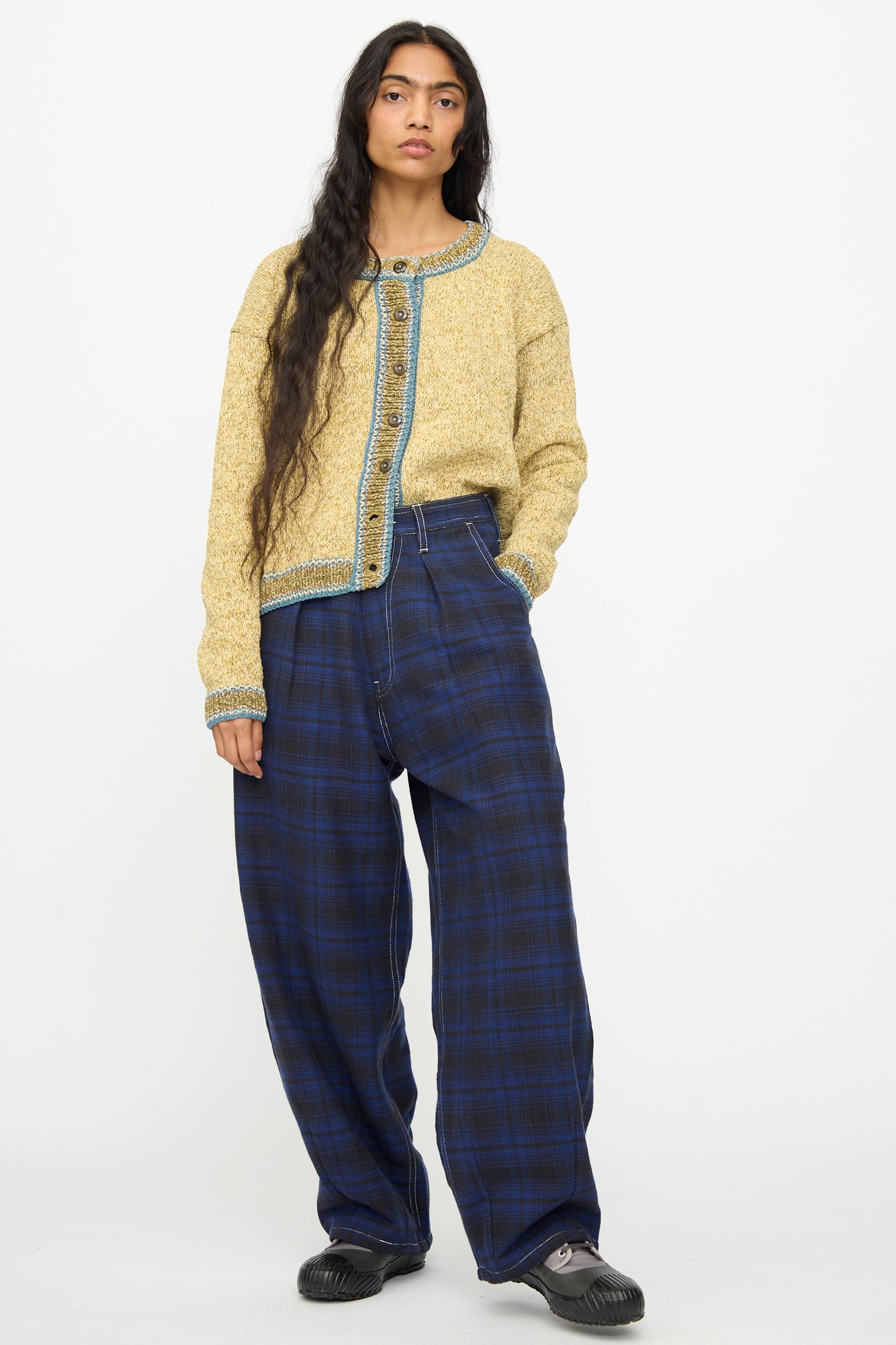 A person wears Story mfg.'s Hand Woven Lush Pants in Solar Check with a textured yellow cardigan featuring blue trim and black shoes, standing against a plain white background.