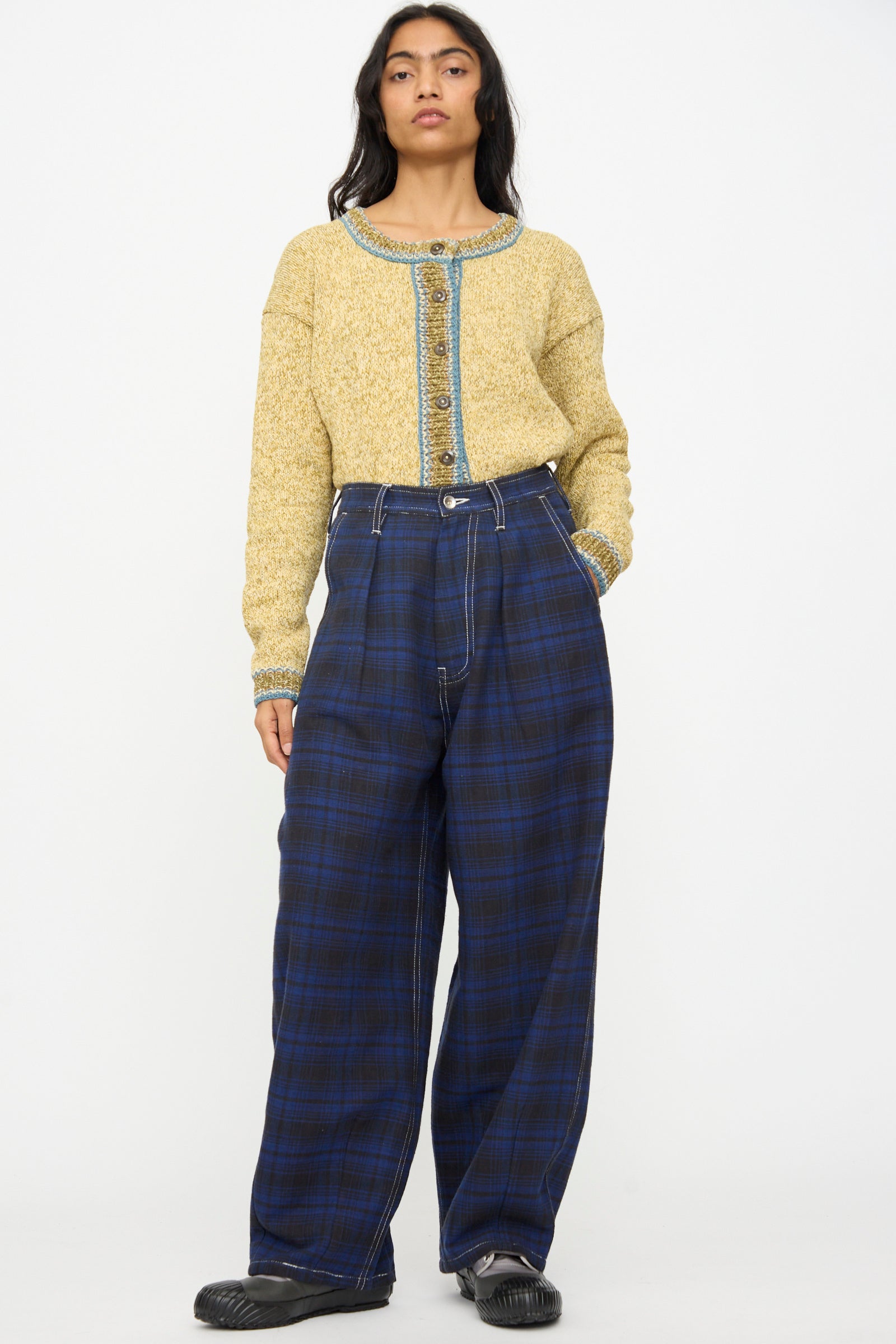 A person wears Story mfg.'s Hand Woven Lush Pants in Solar Check with a textured yellow cardigan featuring blue trim and buttons, paired with black shoes, all set against a plain white background.