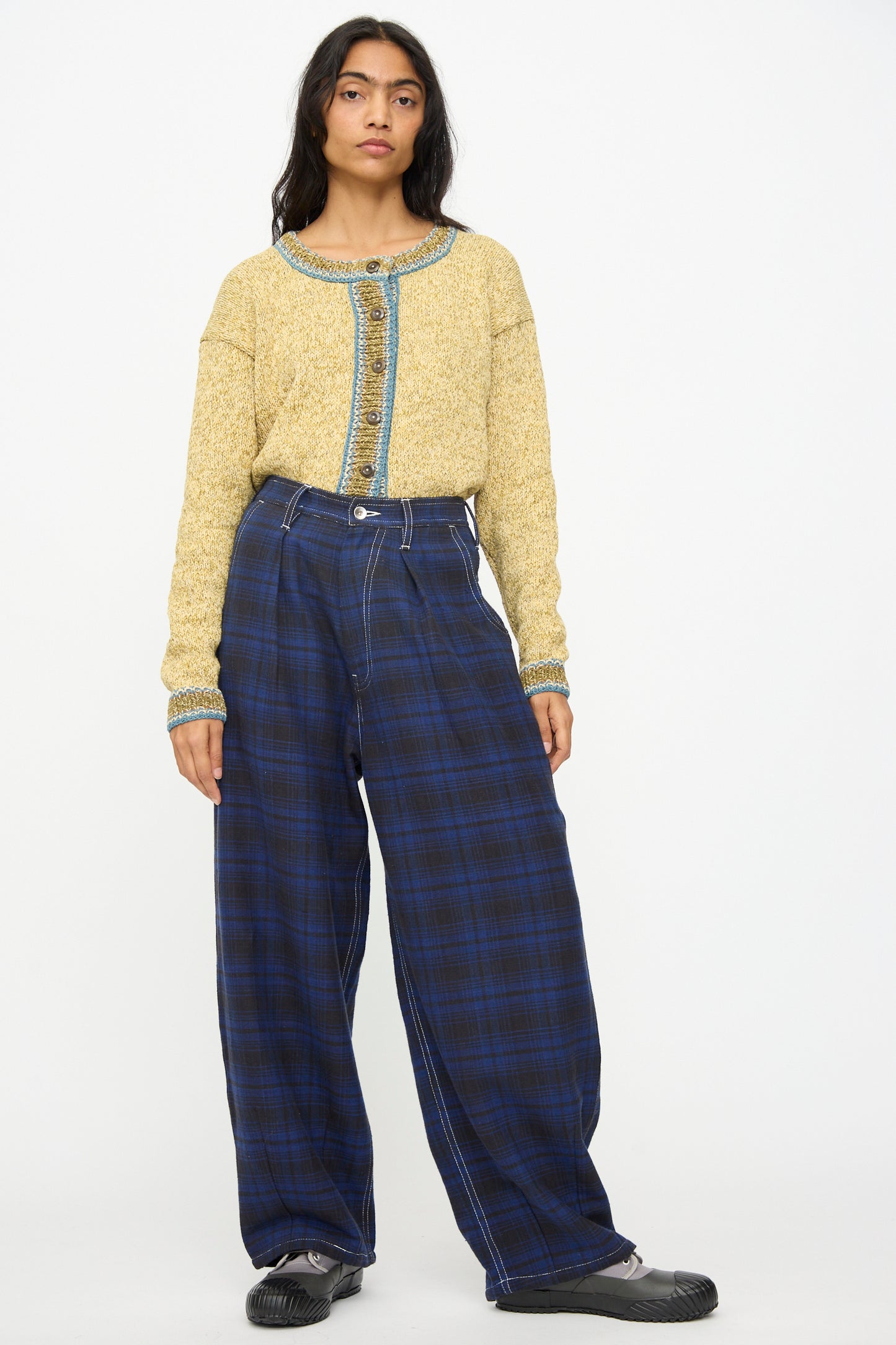 A person wears Story mfg.'s Hand Woven Lush Pants in Solar Check, styled with a yellow knit sweater with blue trim and black shoes, posing against a white background.