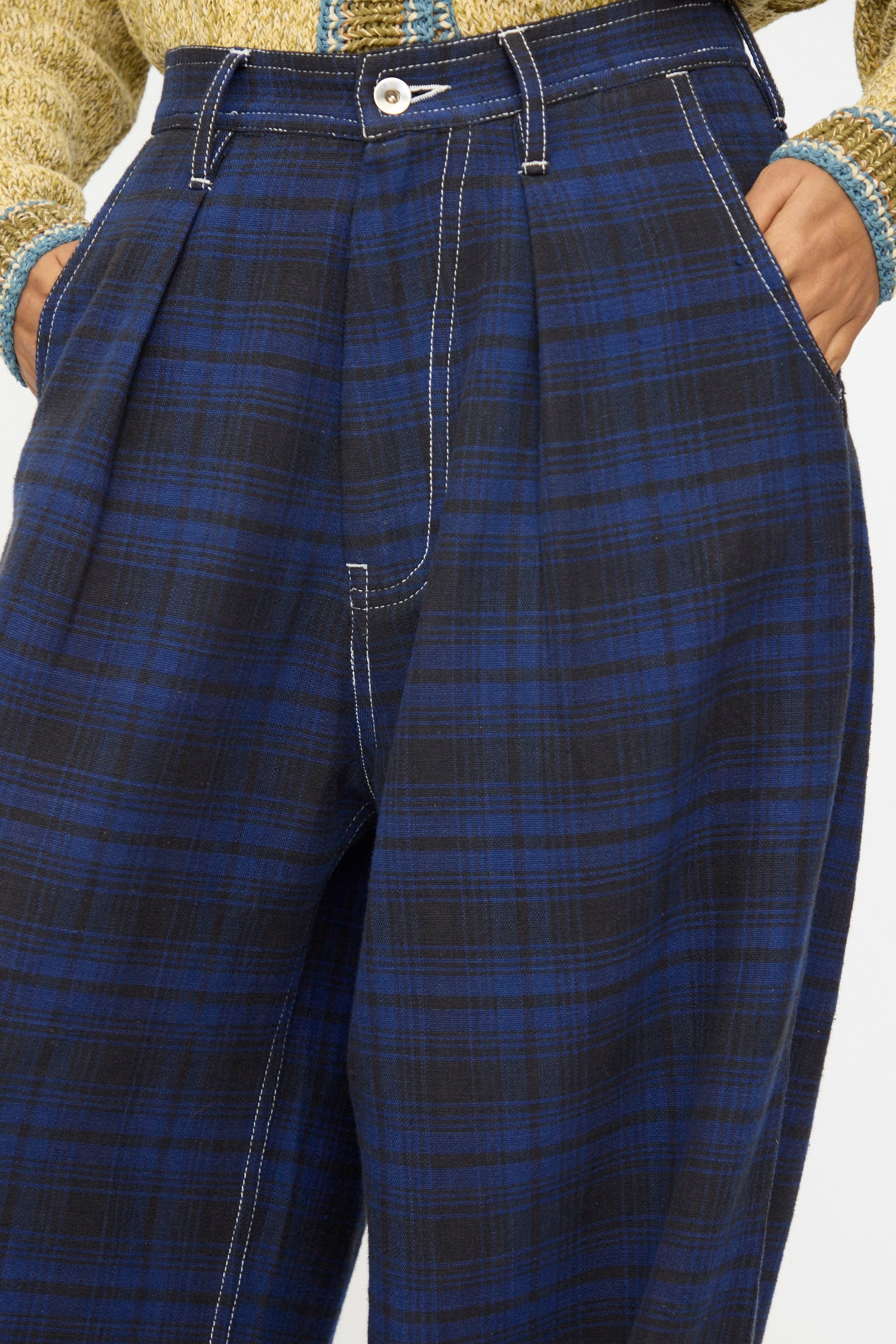Wearing Story mfg.'s Hand Woven Lush Pants in Solar Check—high-waisted blue and black plaid with white stitching—paired with a beige and blue textured sweater, hands in pockets.