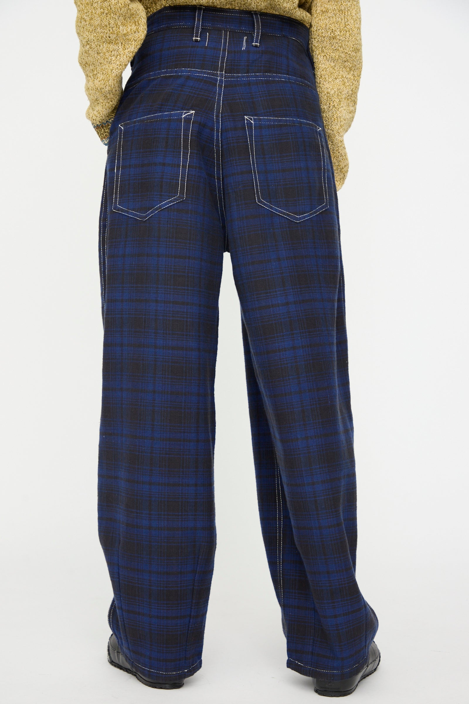 Model wearing Story mfg. Hand Woven Lush Pants in Solar Check—blue and black plaid with visible white stitching—and a beige sweater, shown from the back against a plain background.