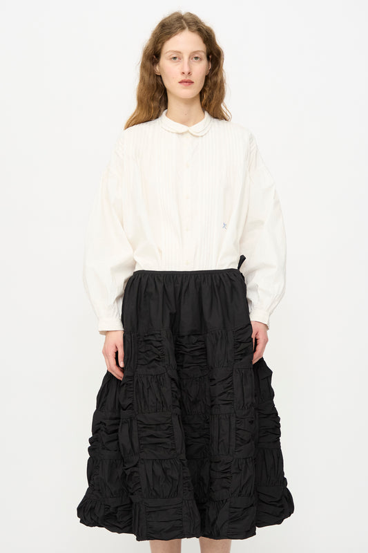A person with long wavy hair wears a white button-up shirt and the Story mfg. Patchwork Foxglove Skirt in Interstellar, standing against a plain white background.