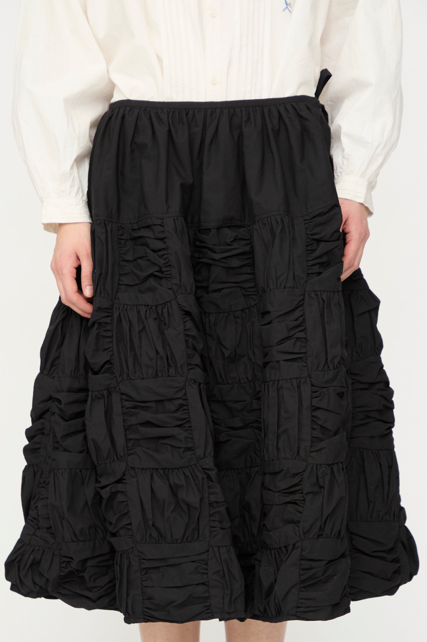 A person wearing a white long-sleeve shirt and the Story mfg. Patchwork Foxglove Skirt in Interstellar, a black checker patchwork, knee-length skirt with gathered square panels.
