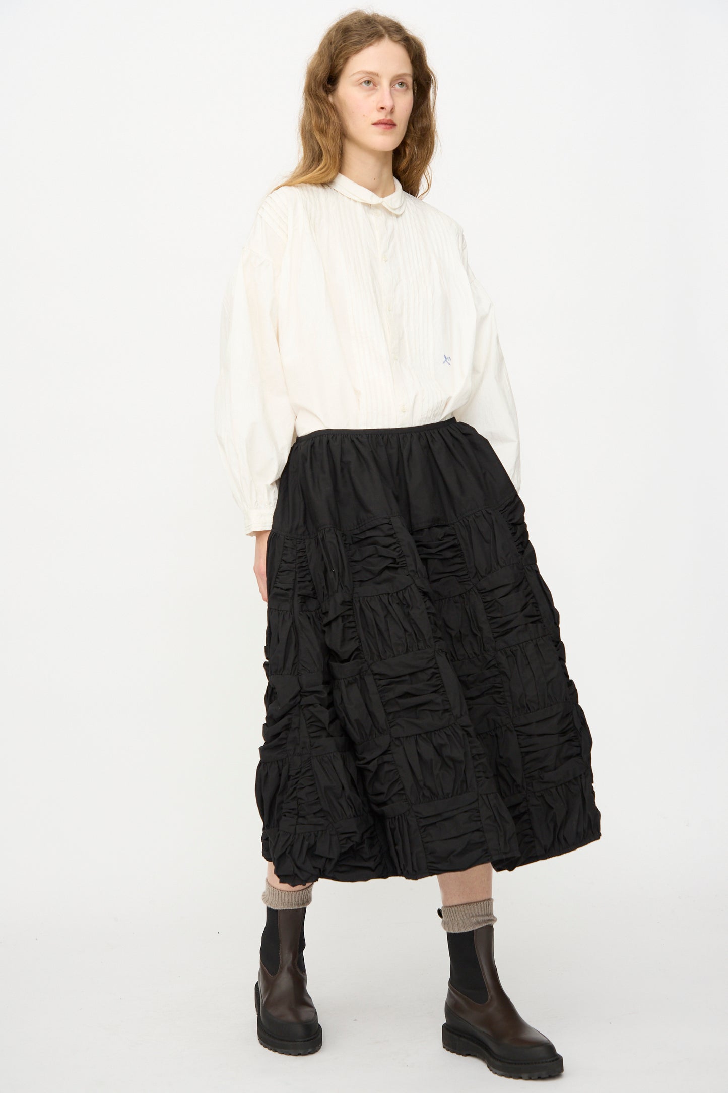 A person stands against a plain white background wearing the Story mfg. Patchwork Foxglove Skirt in Interstellar, paired with a loose white blouse, beige socks, and dark ankle boots.